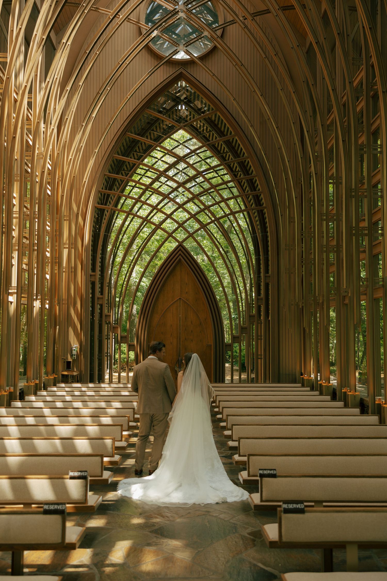Bride and groom walk down the aisle of a stunning wood and glass Gothic chapel surrounded by lush green trees.