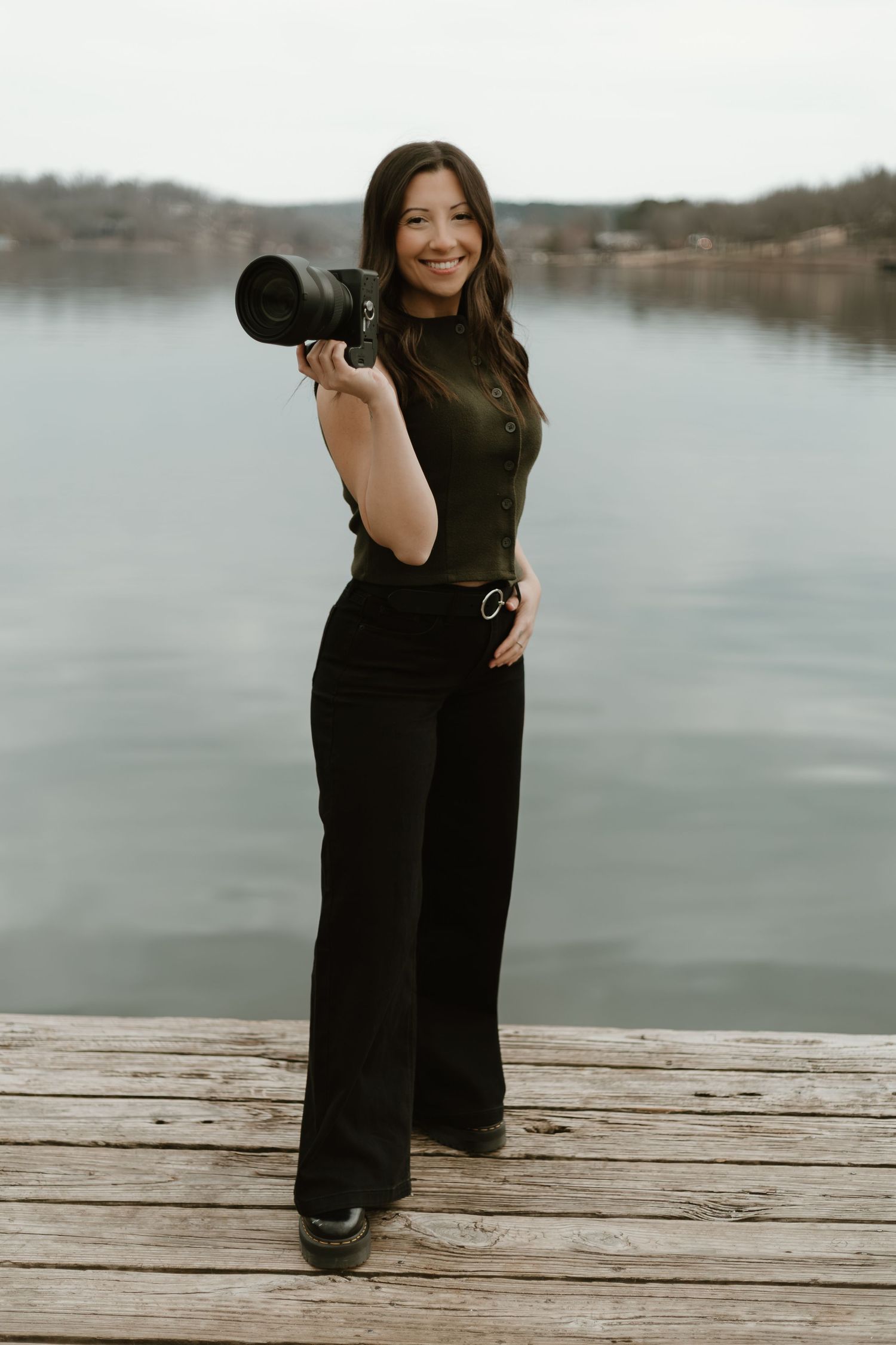 A photographer in a black dress poses with a camera on a wooden dock overlooking a serene lake.