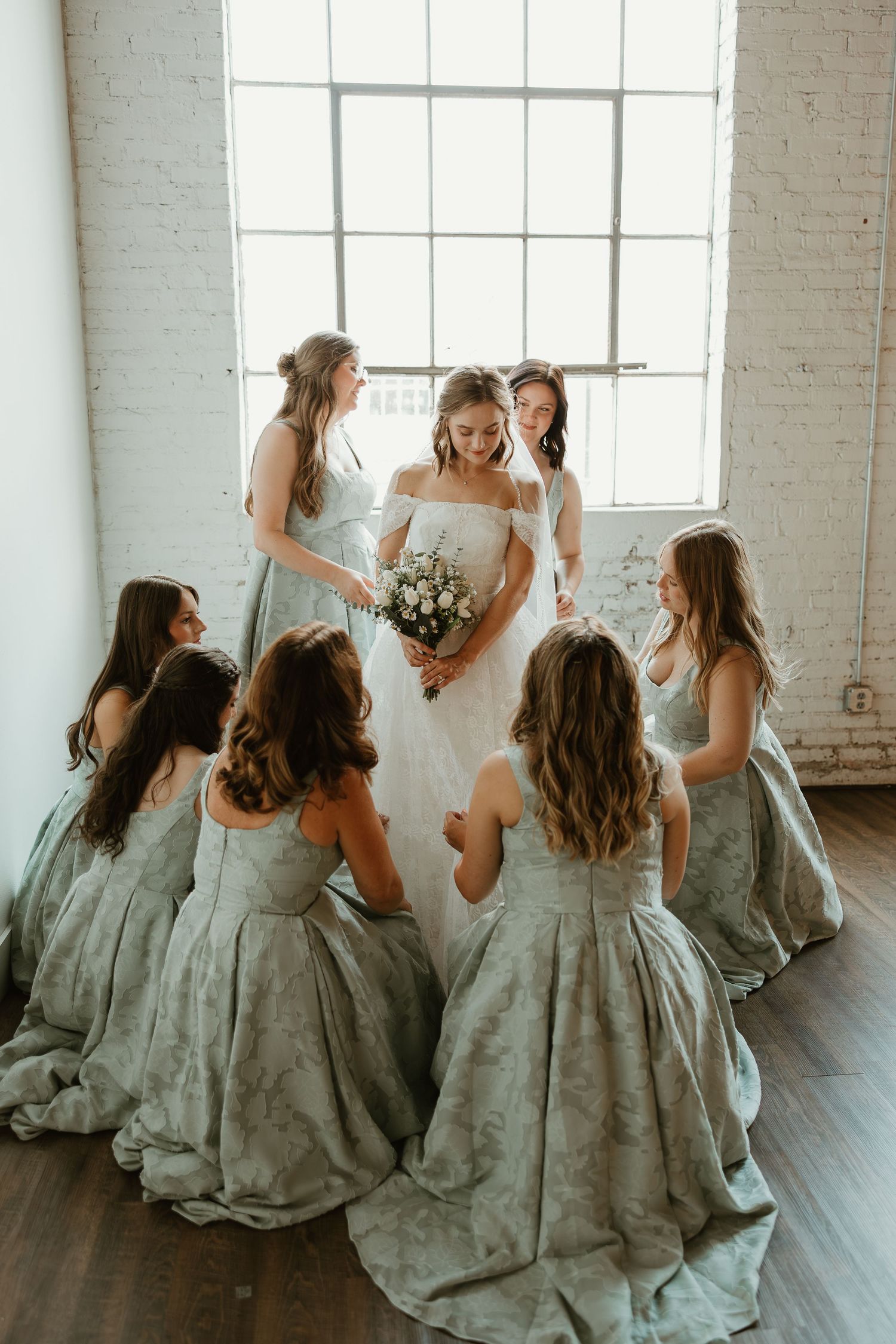 A wedding party in matching gray dresses laughs and dances together in a bright white studio space.