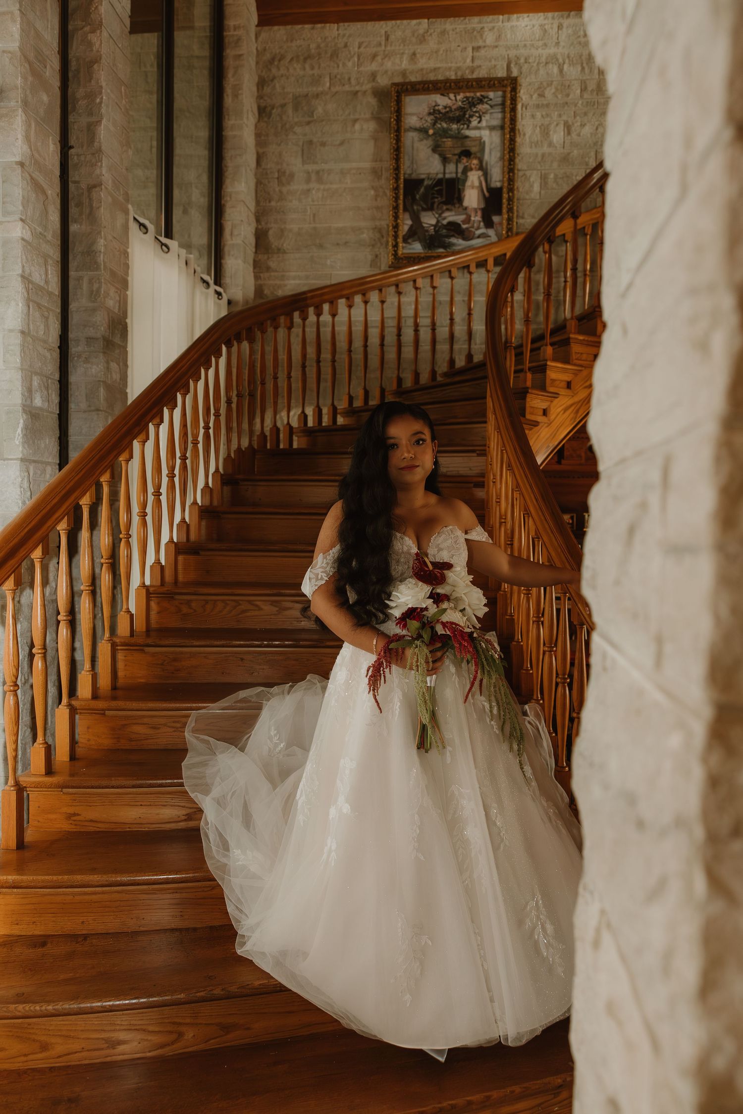 A bride in a flowing white gown poses gracefully on a grand wooden staircase with brick walls in the background.