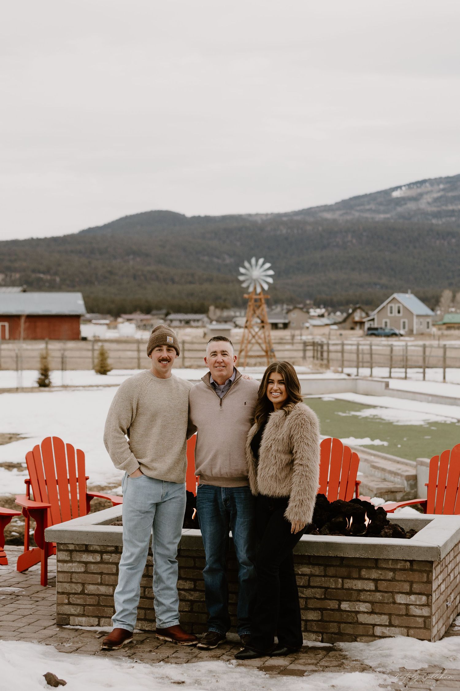 A winter mountain scene with red Adirondack chairs and decorative windmill by a stone fire pit in a snowy backyard.