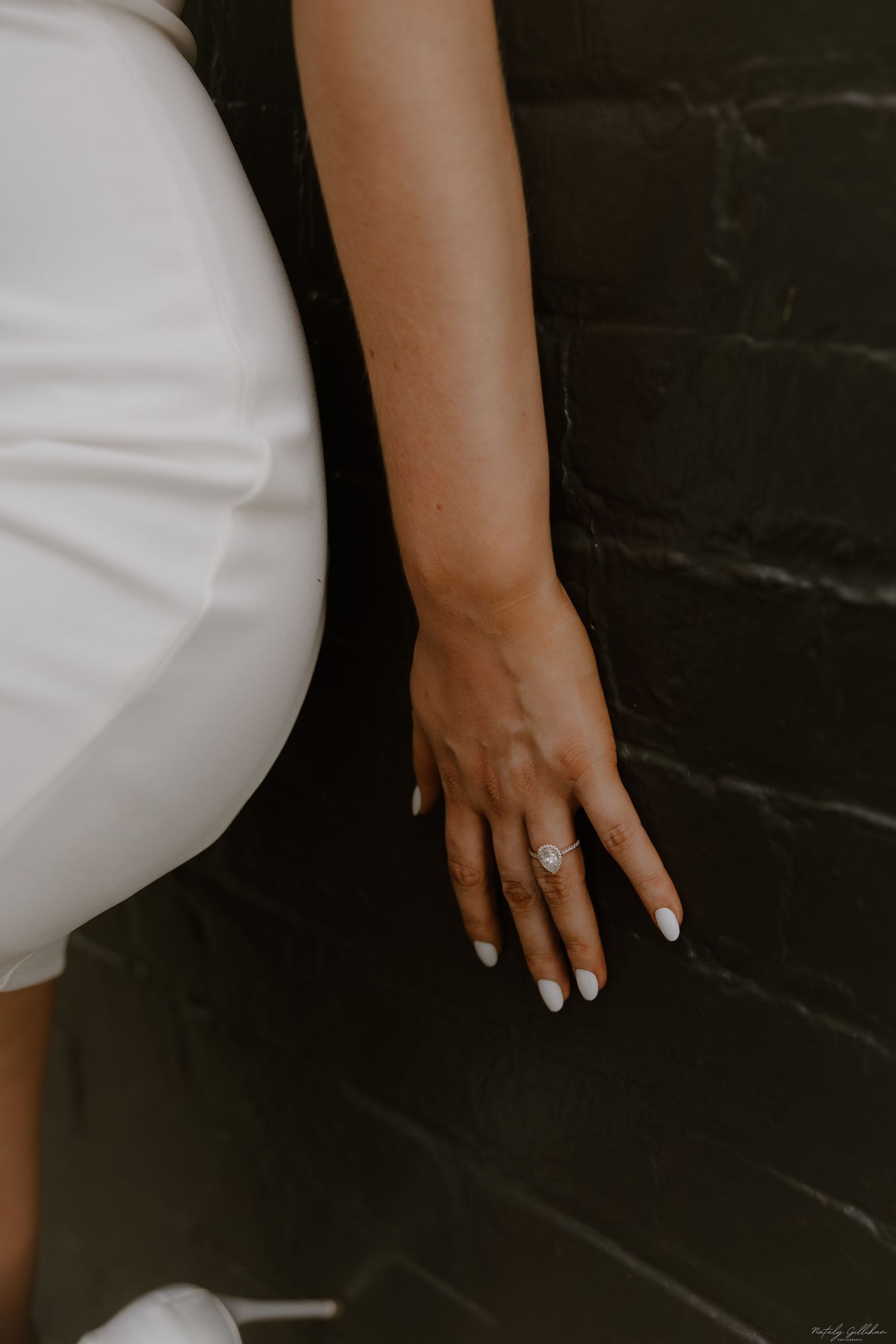 Close-up of an elegant hand with white nail polish and engagement ring against a dark wall.