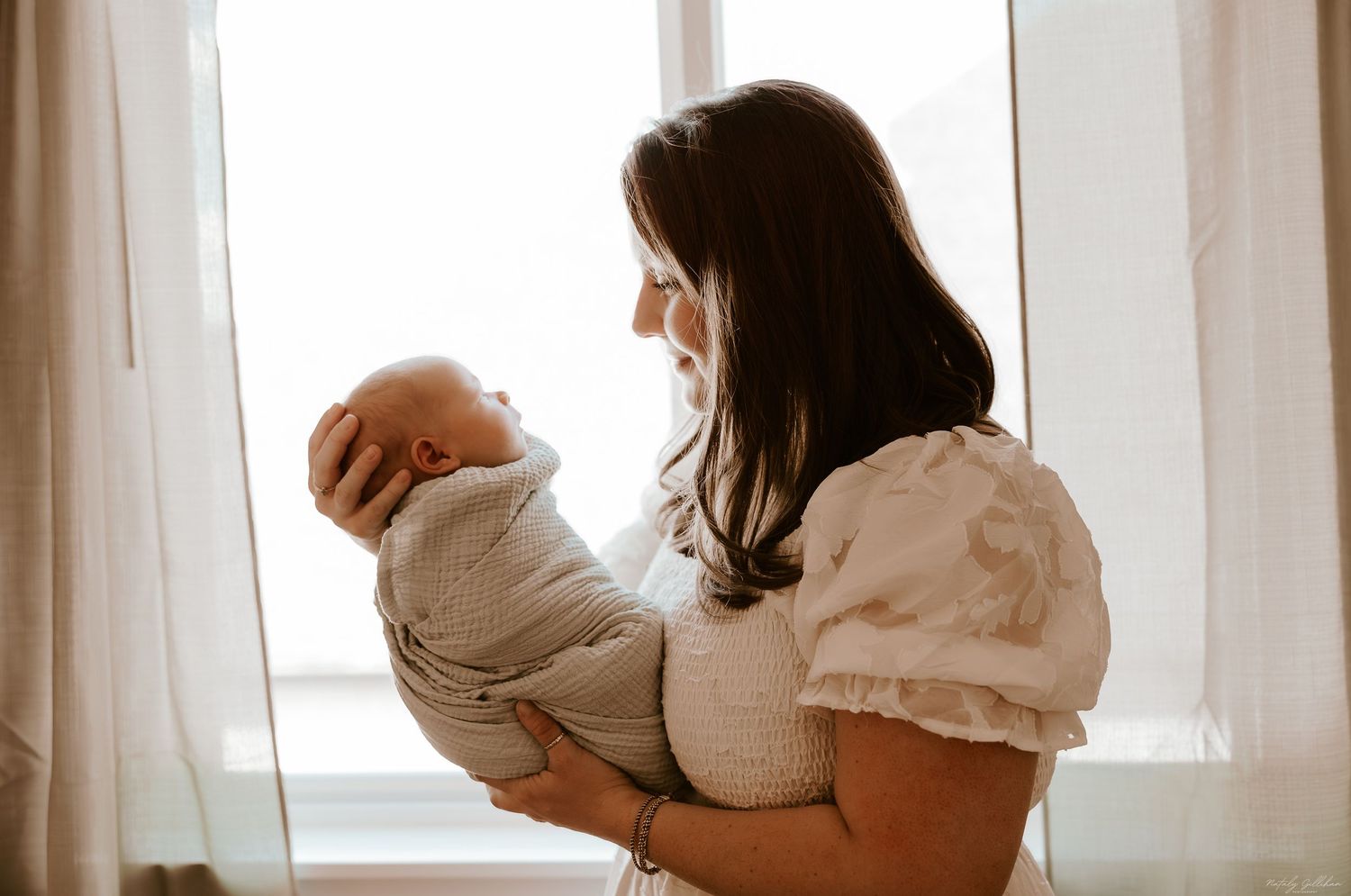 A figure in cream-colored clothing holds a baby while standing near a sunlit window with sheer curtains.