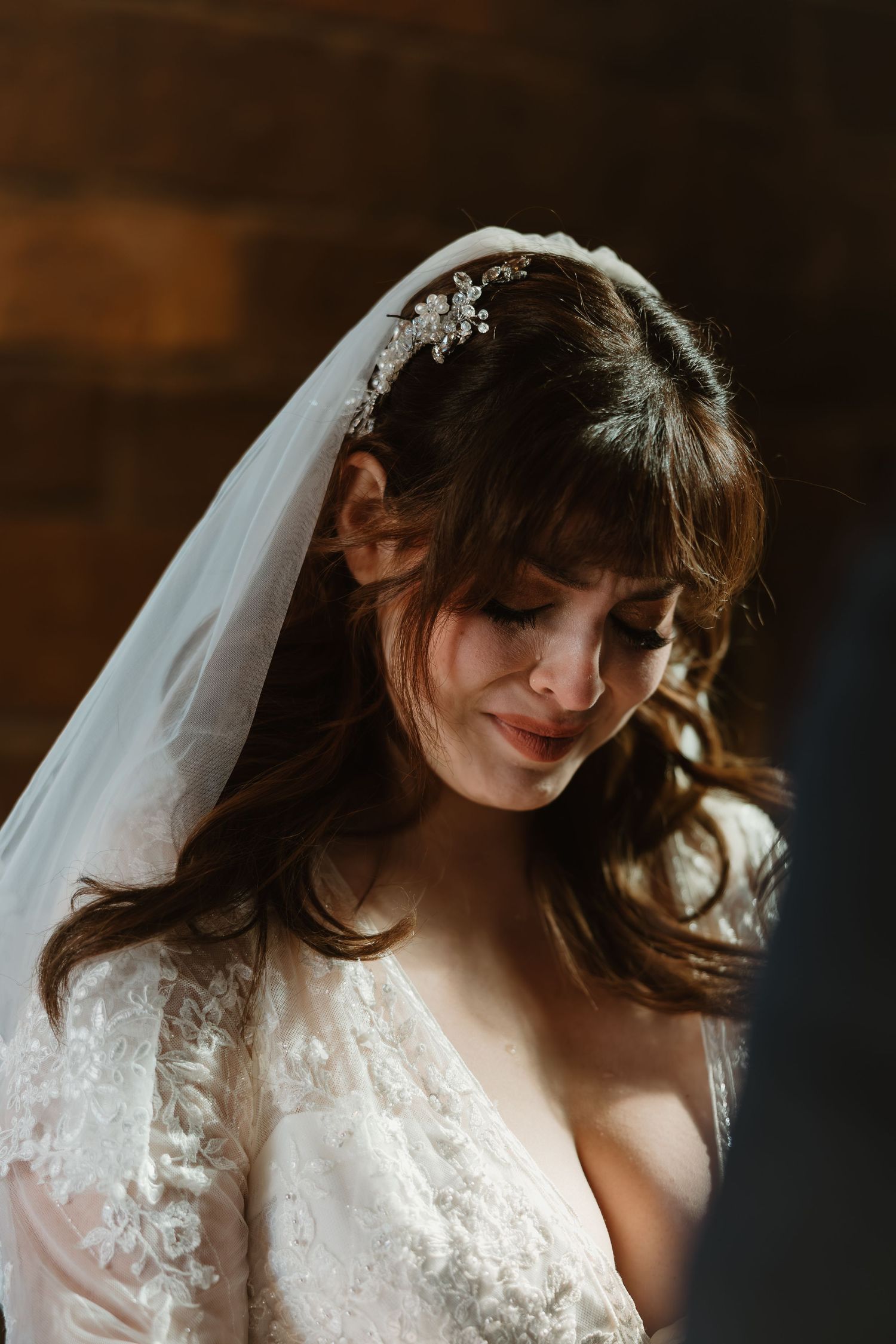 A bride in a lace wedding dress with flowing veil poses in soft lighting against a dark background.