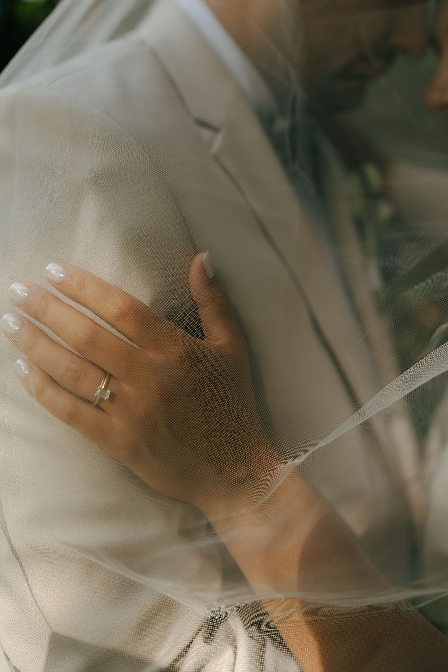 Bride's hand with gold wedding ring resting on groom's white suit jacket, partially covered by a sheer veil.