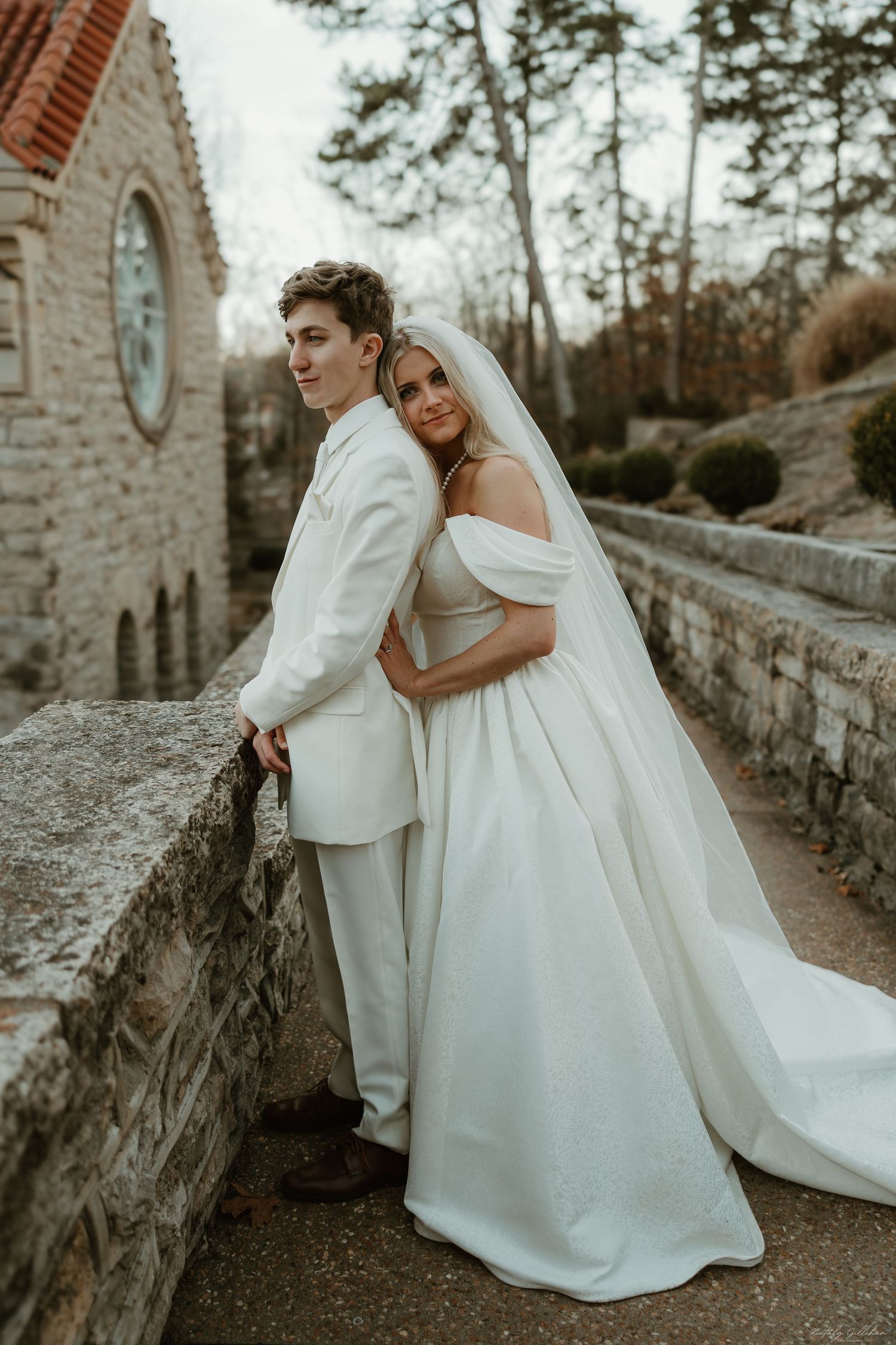 A bride and groom in matching white attire pose together against a stone wall at a romantic outdoor venue.