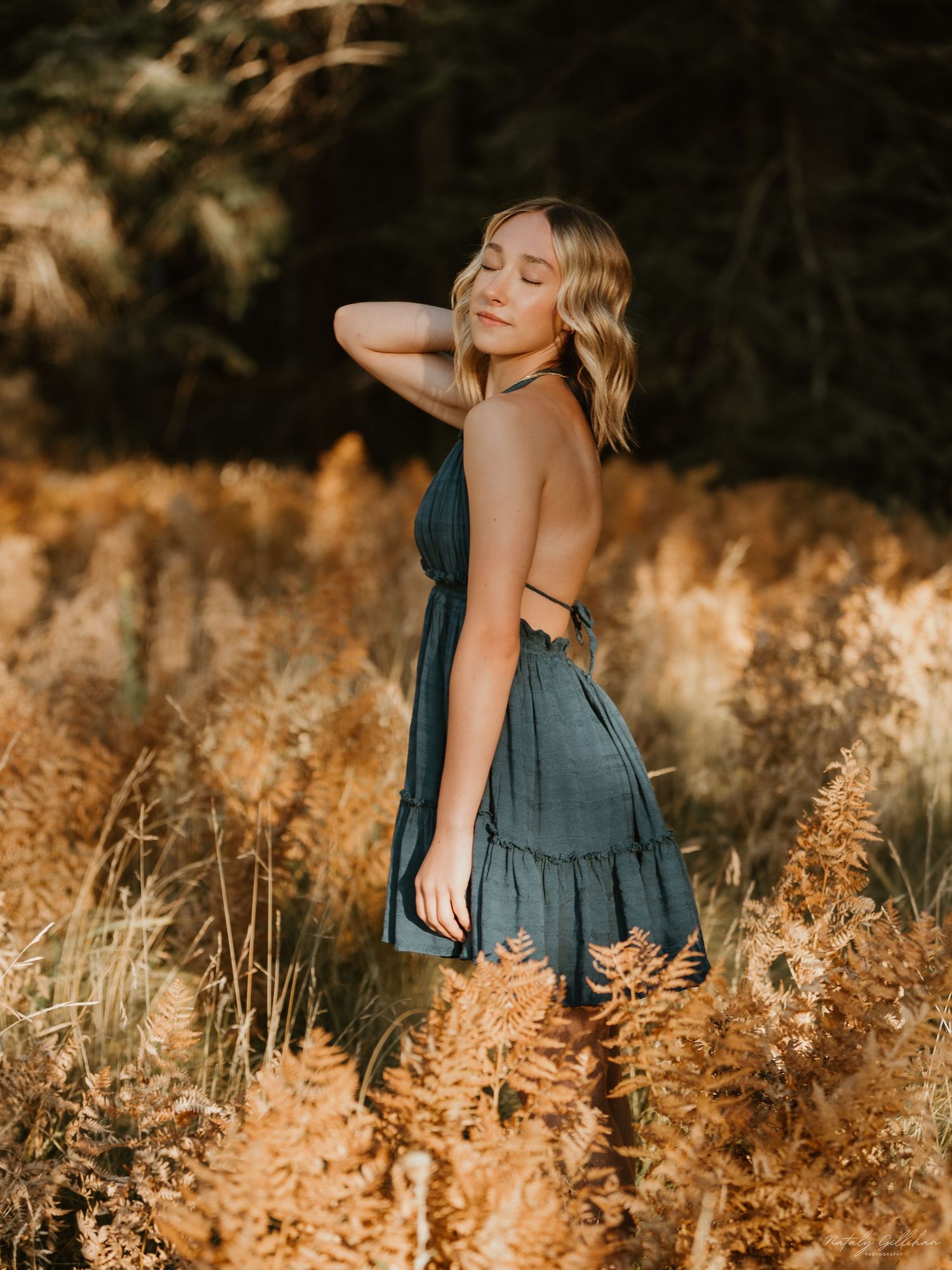 Person in a blue sun dress poses gracefully among golden tall grass during sunset lighting in an outdoor setting.