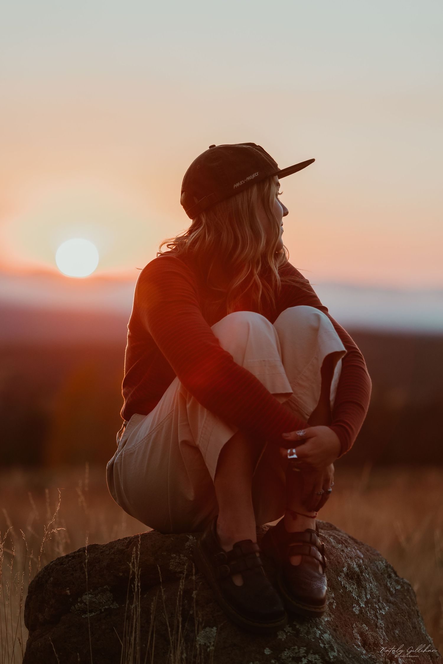 Silhouetted figure in baseball cap sits on rock watching colorful sunset over mountain landscape.