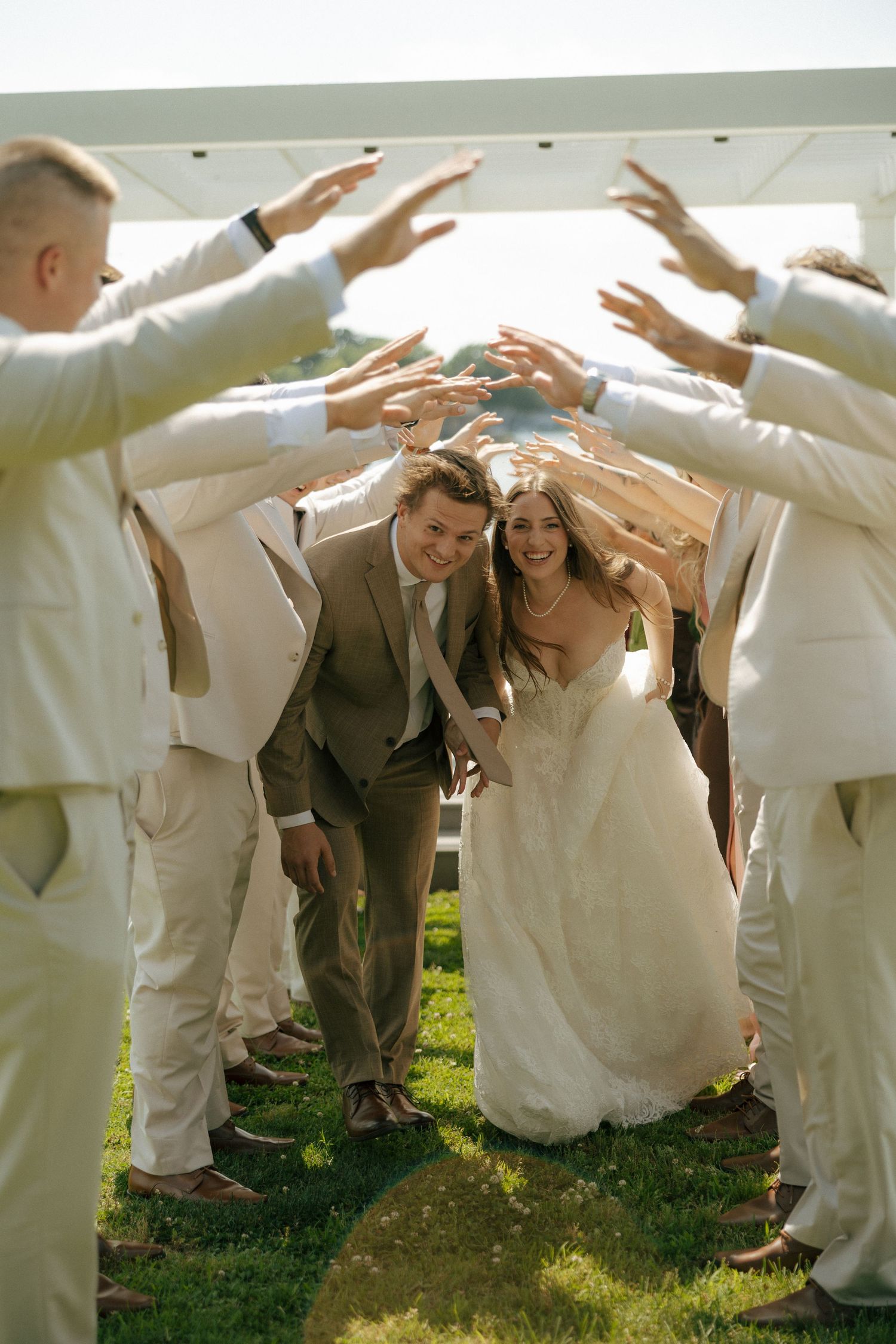 Bride and groom walk through an arch of raised hands formed by wedding party on sunny green lawn.