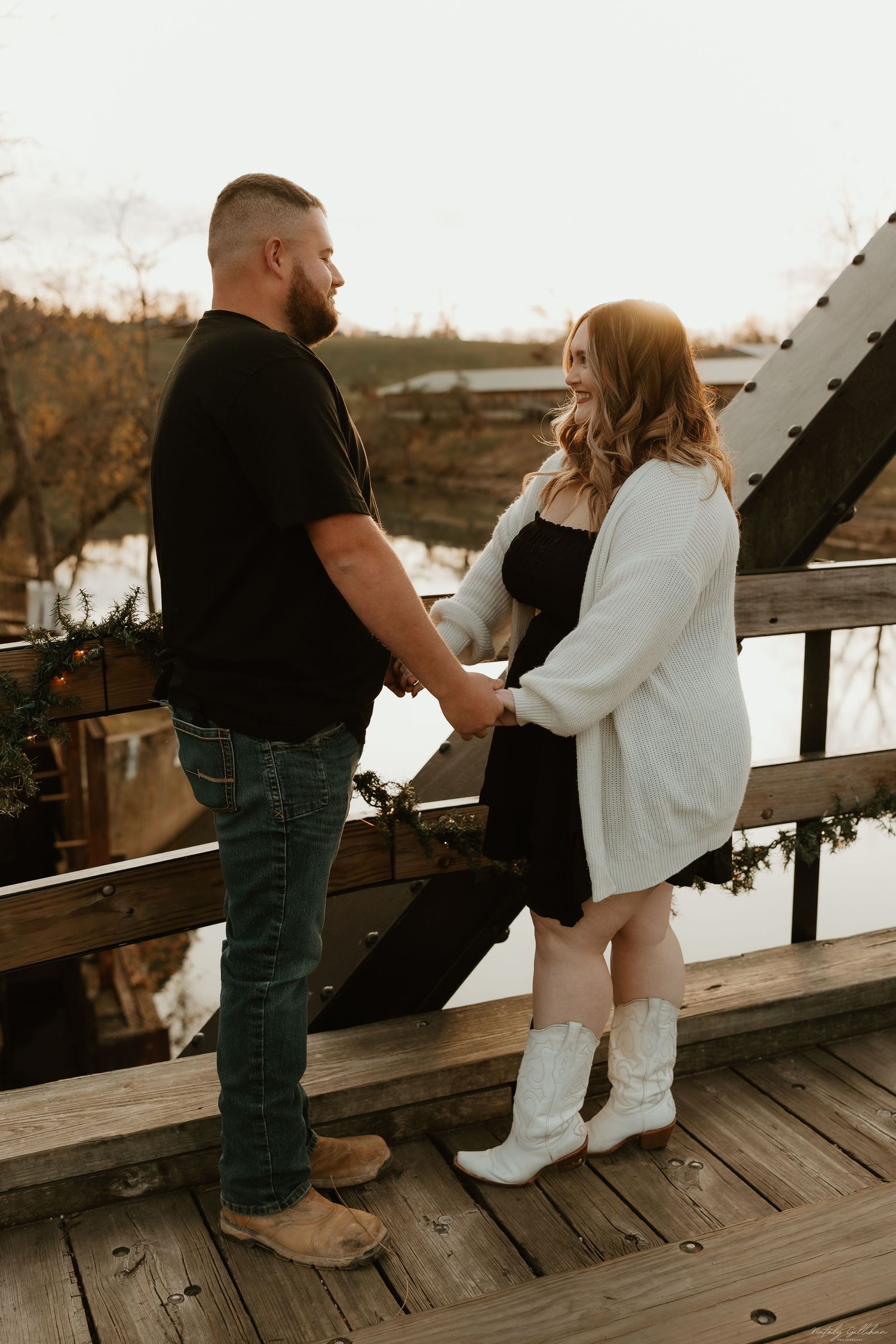 A couple stands holding hands on a rustic wooden bridge during a warm autumn sunset.