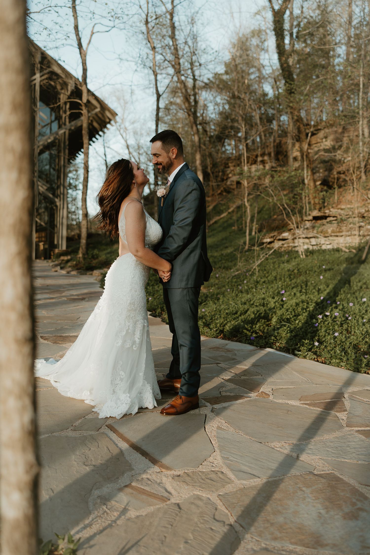 A bride in a white lace wedding gown and groom in gray suit embrace on a stone patio surrounded by autumn trees.