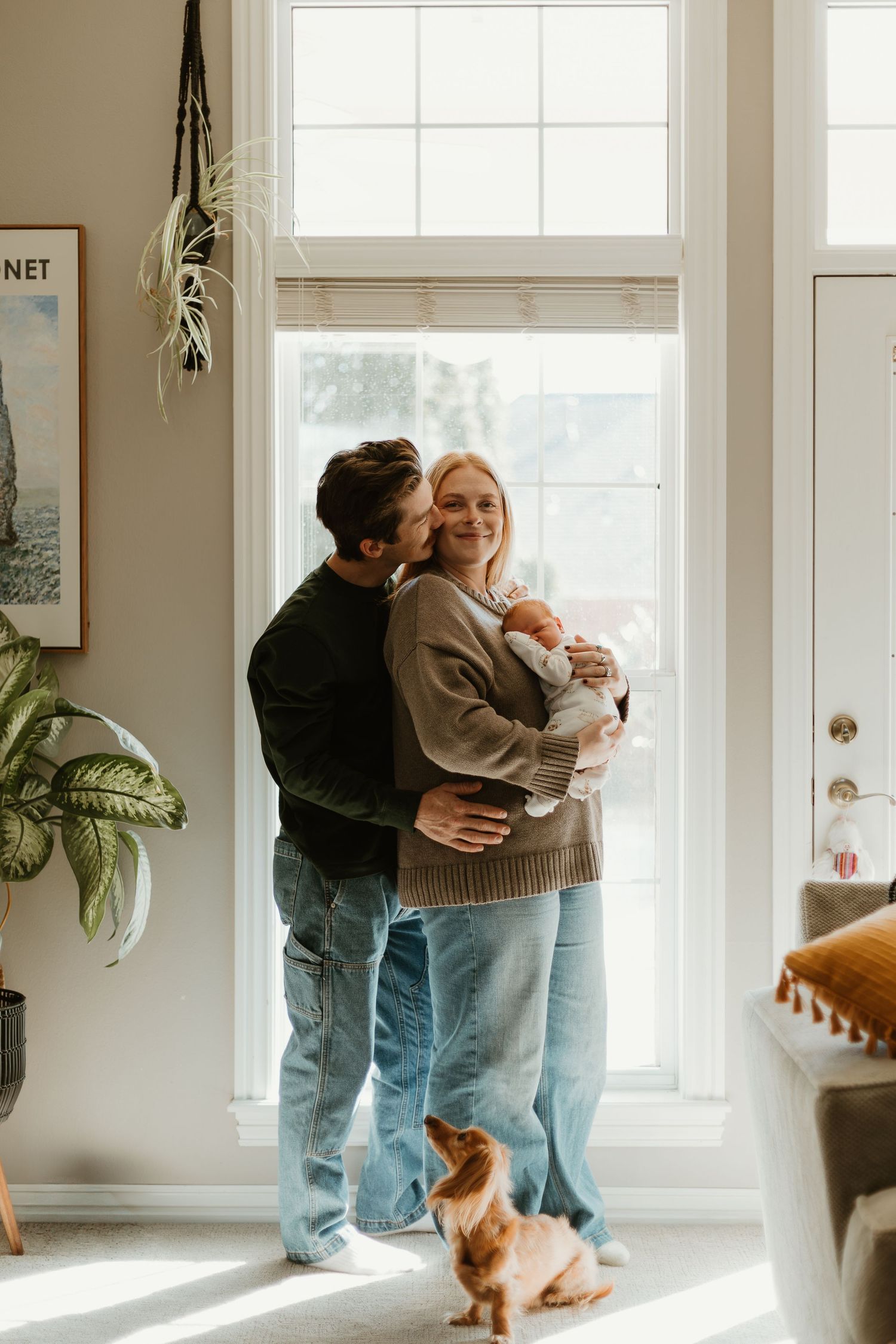 A cozy indoor scene with people embracing near bright windows while a ginger cat watches from the floor.