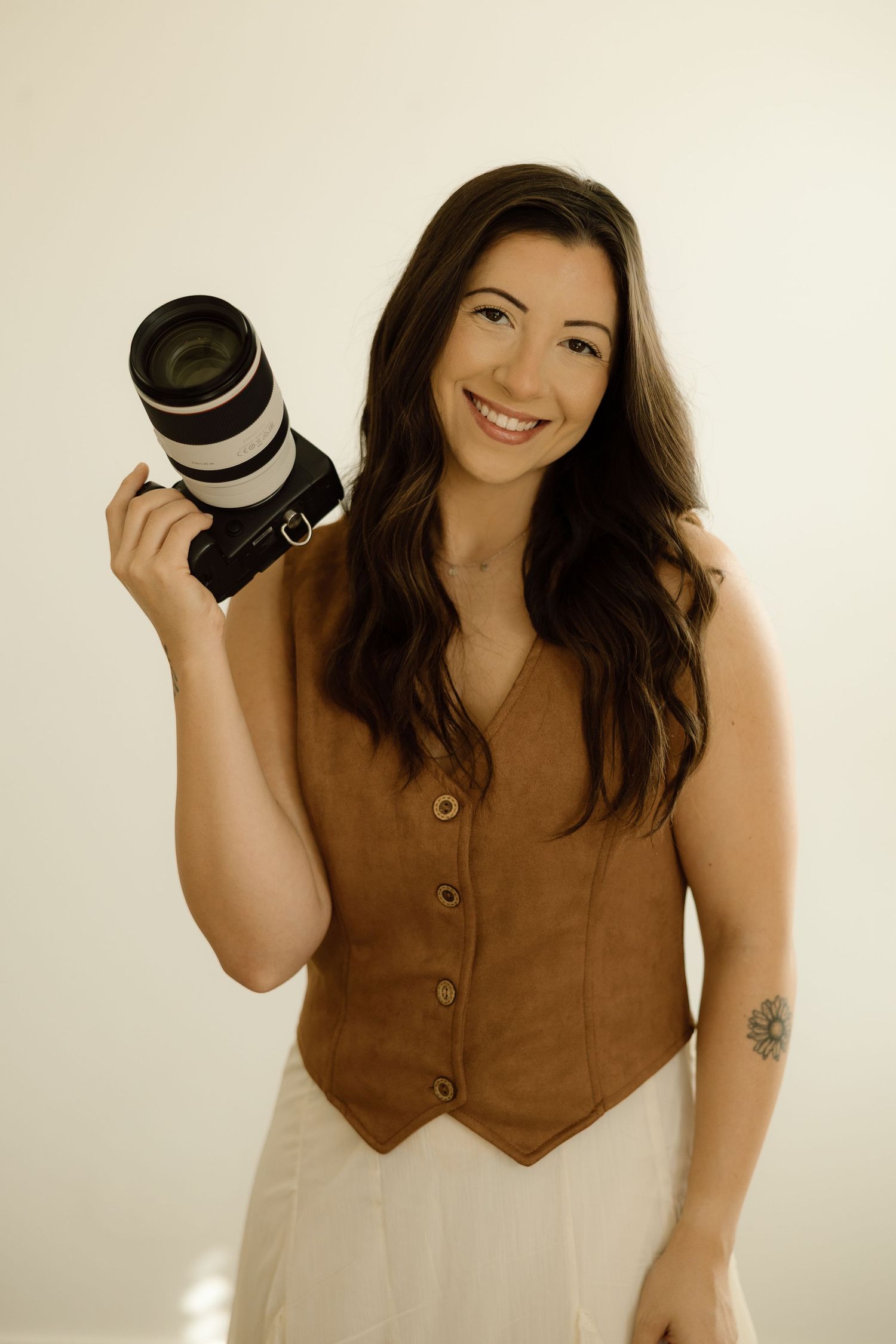 Smiling woman photographer holding a Canon telephoto lens camera, wearing a brown vest and white skirt with a floral tattoo.