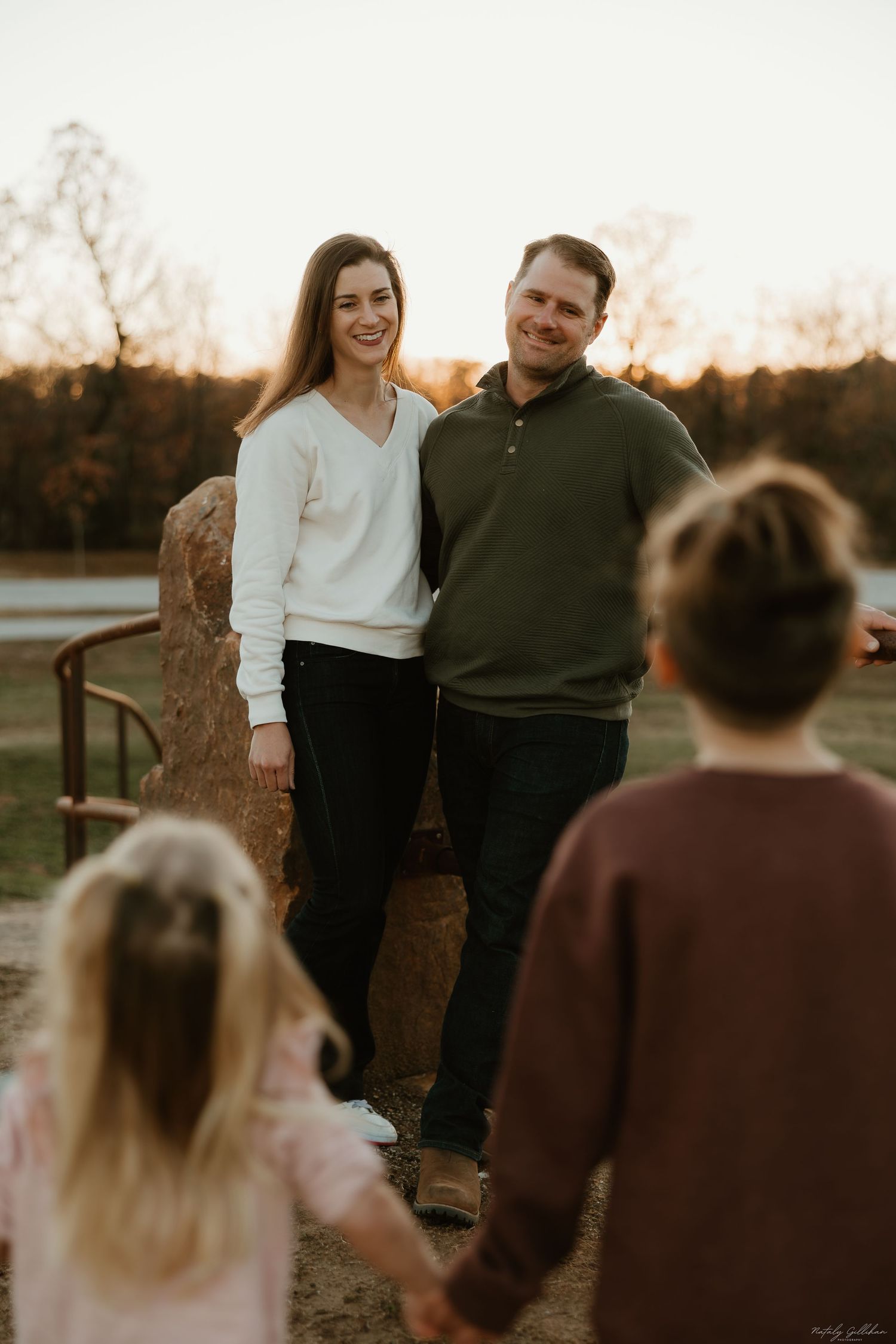 A couple stands together at sunset while being photographed outdoors in warm golden light.