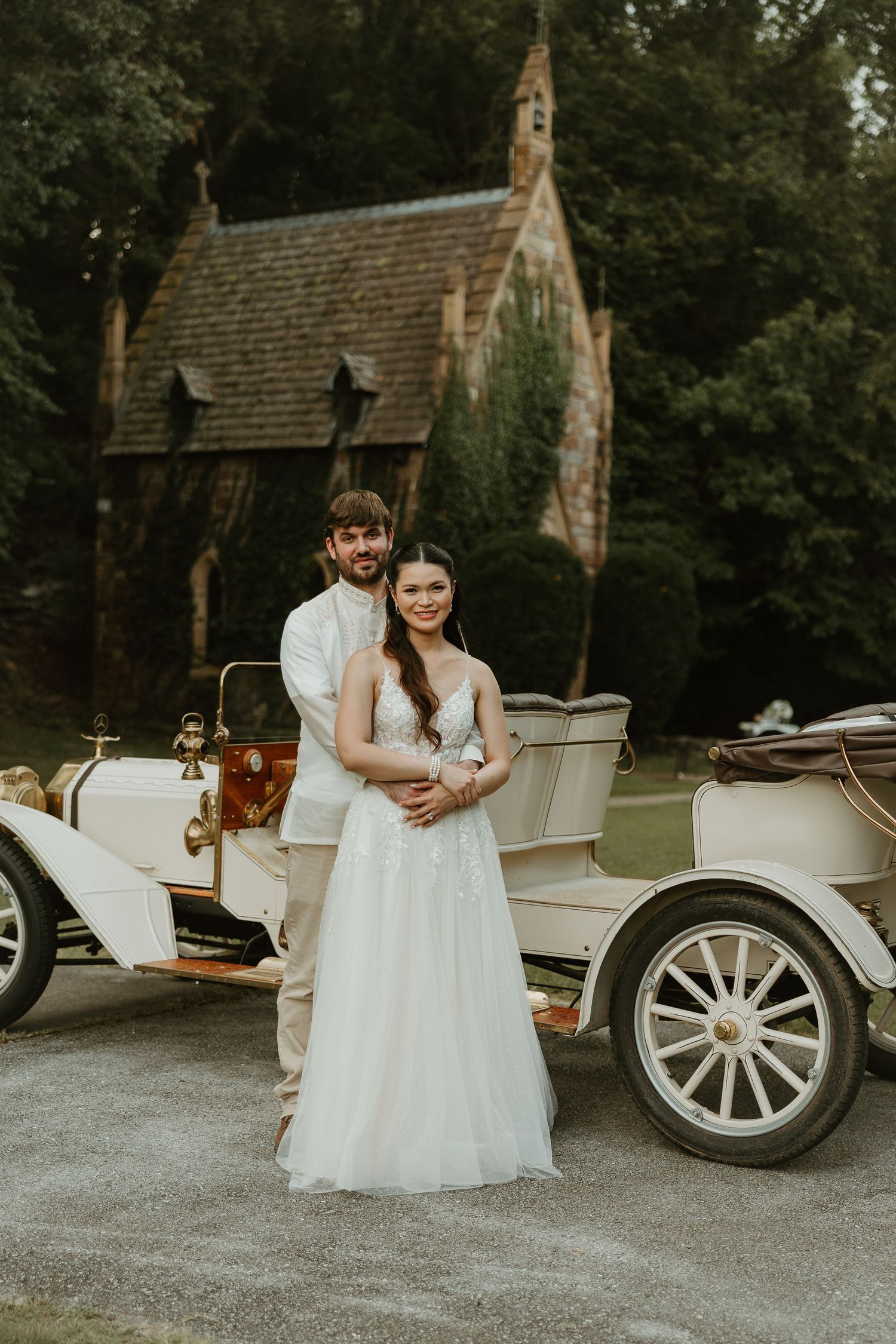 Romantic couple in white attire sitting together near vintage carriage at outdoor wedding venue.