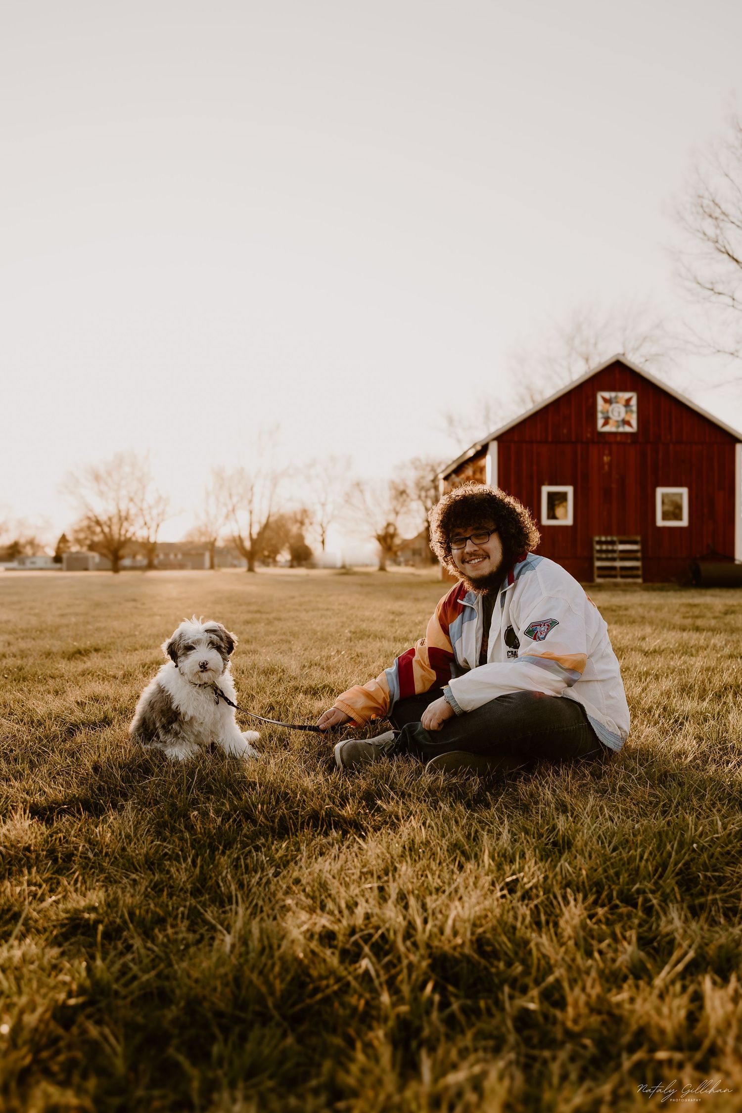 Person and Australian Shepherd dog sitting in grass at sunset with red barn in background on autumn evening.