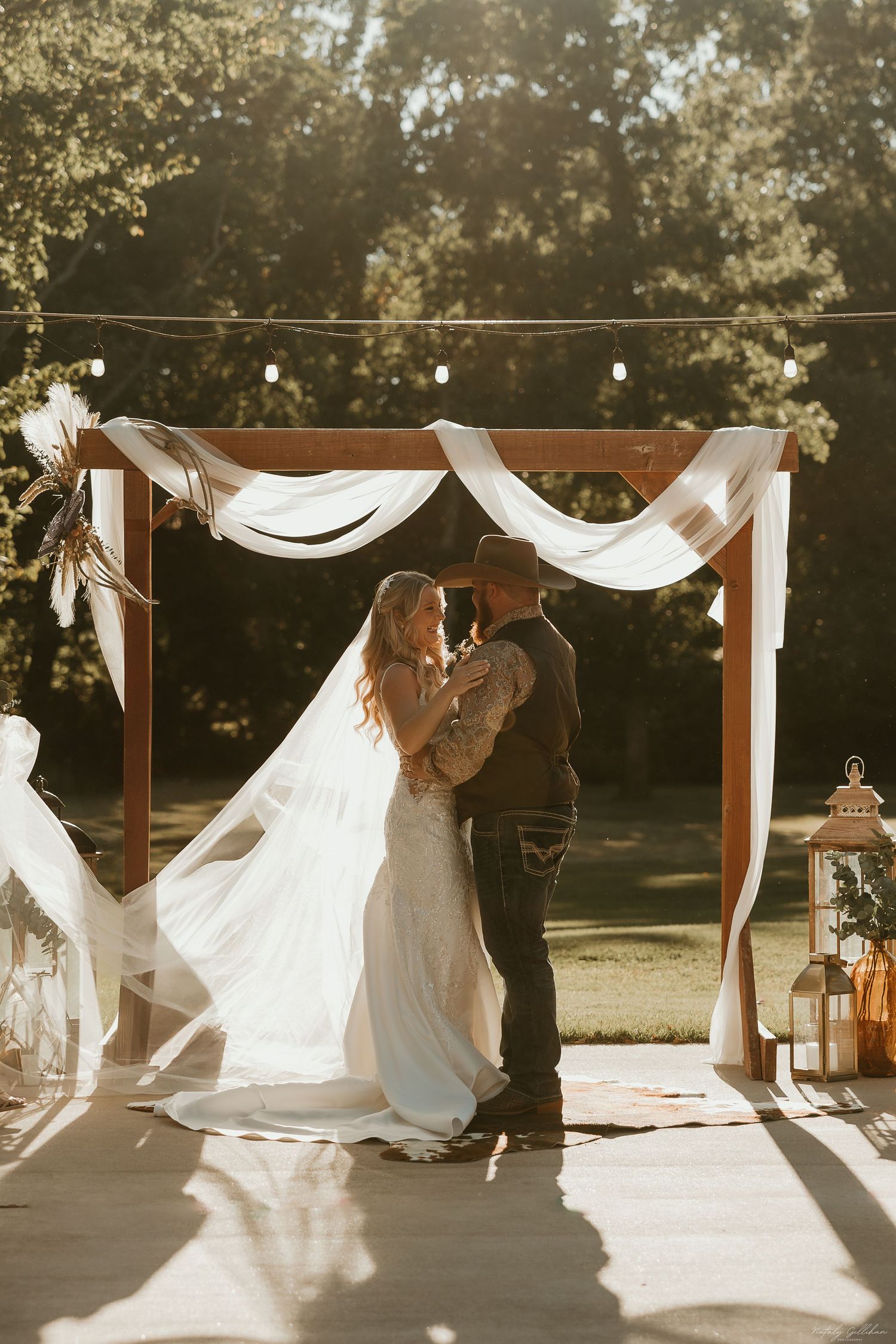 Couple embraces under wooden wedding arch decorated with white fabric and lanterns at sunset outdoors.