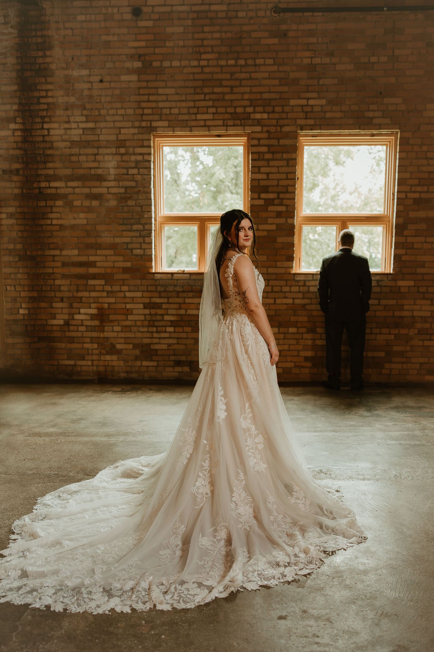 A bride in a lace wedding gown with long train stands in a rustic brick room while a silhouette gazes out the window.