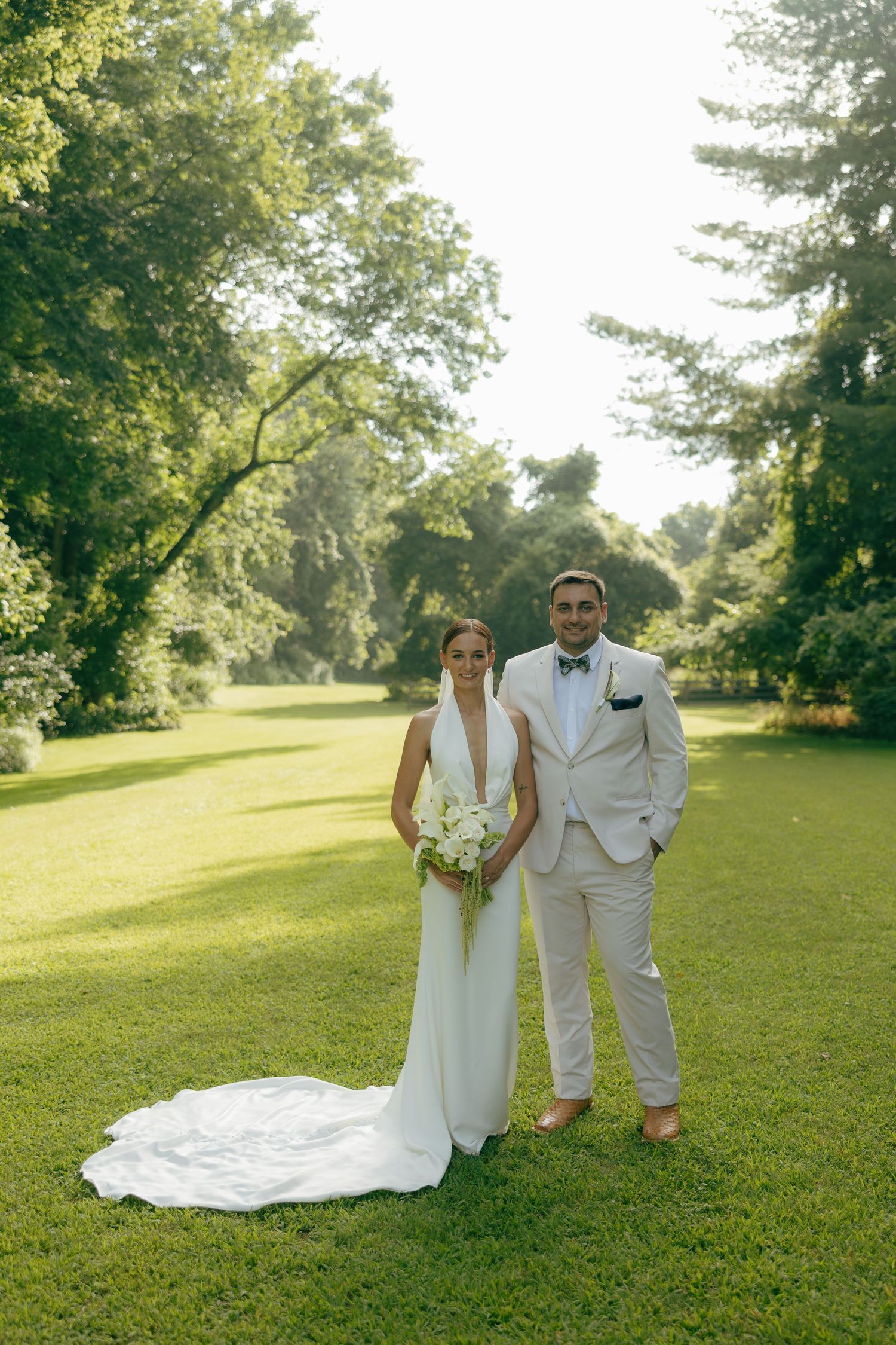 Bride and groom standing on lush green lawn surrounded by trees in elegant white wedding attire with bridal bouquet.