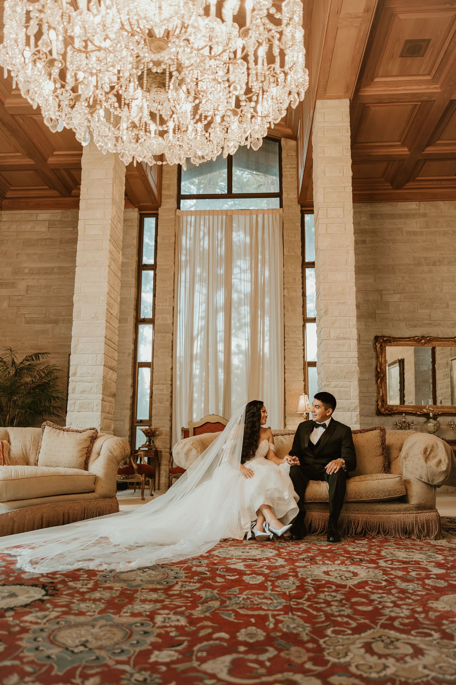 A couple sits beneath an elegant crystal chandelier in a luxurious room with tall windows and ornate red carpet.