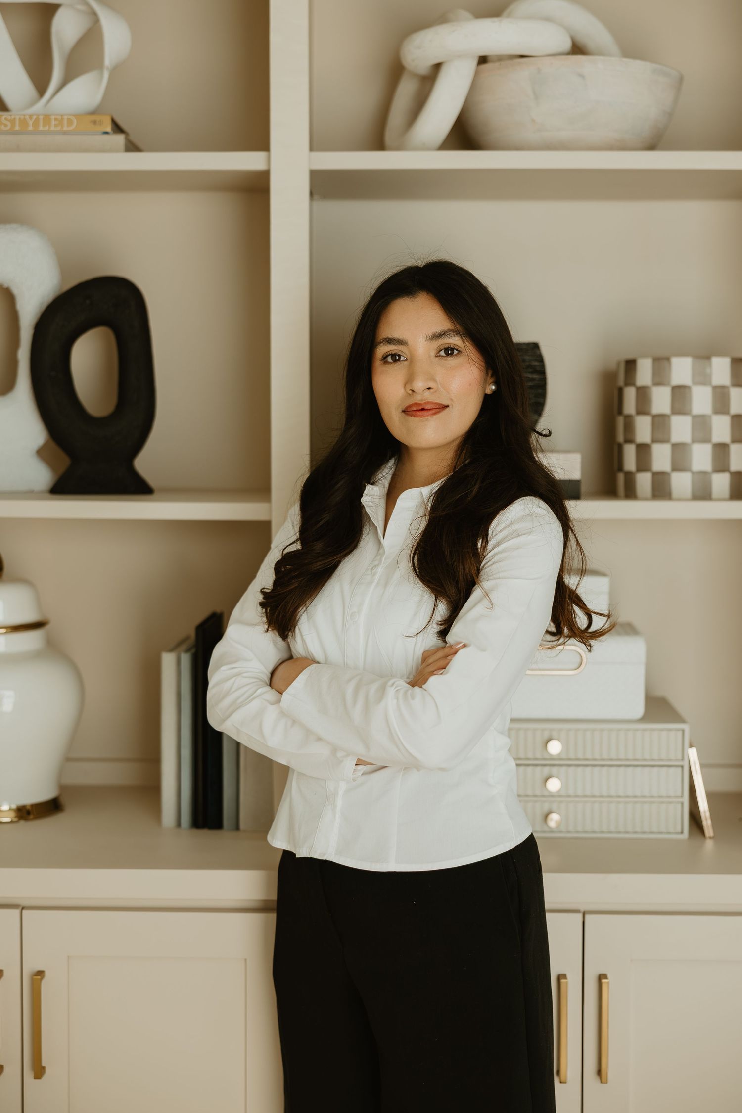 A professional woman in a white blouse stands confidently with arms crossed before a styled beige bookshelf.