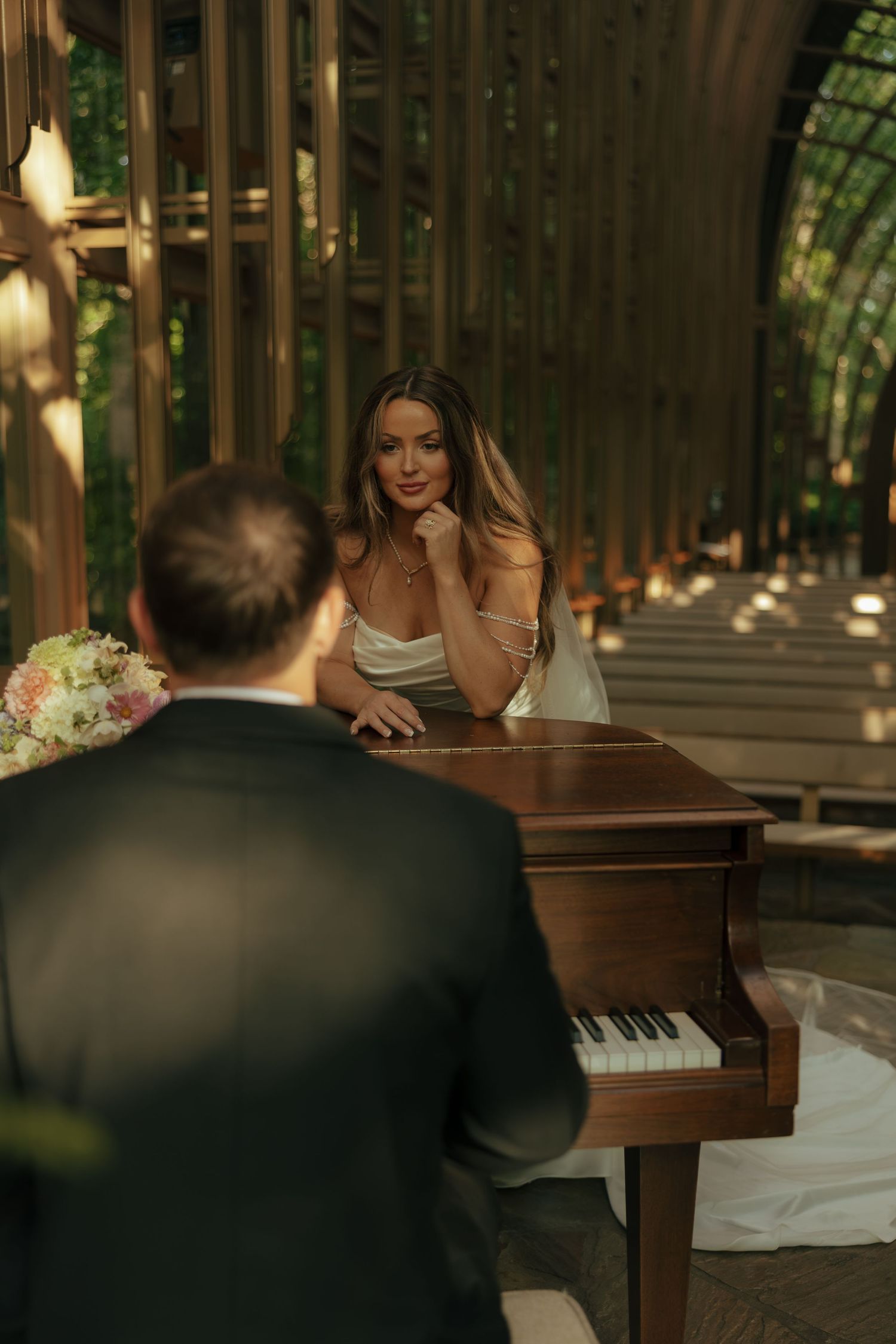 Bride in white dress leans on piano smiling at groom in dark suit during elegant woodland wedding ceremony.