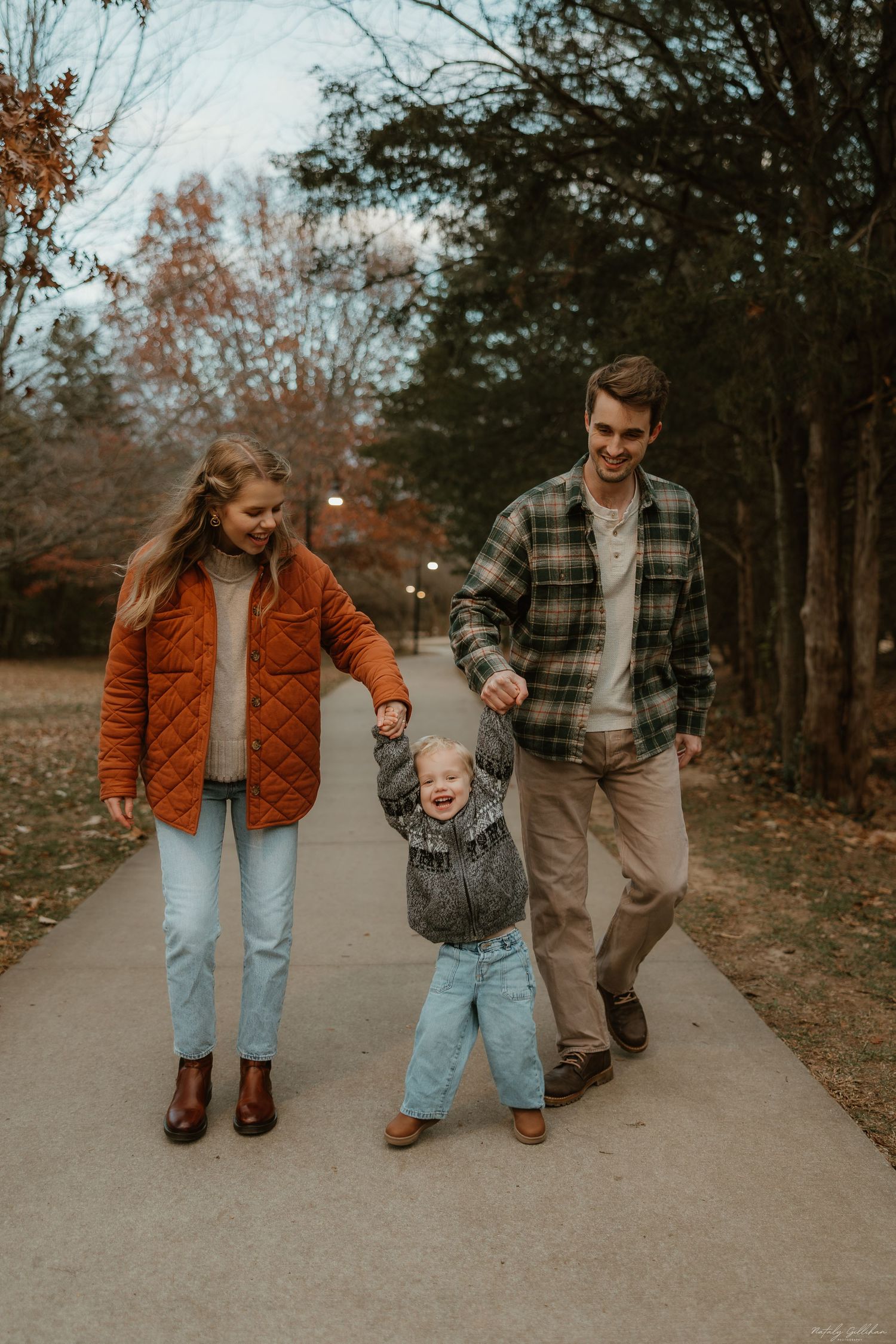 A family walks together along a wooded path on an autumn day, with the little one joyfully swinging between them.