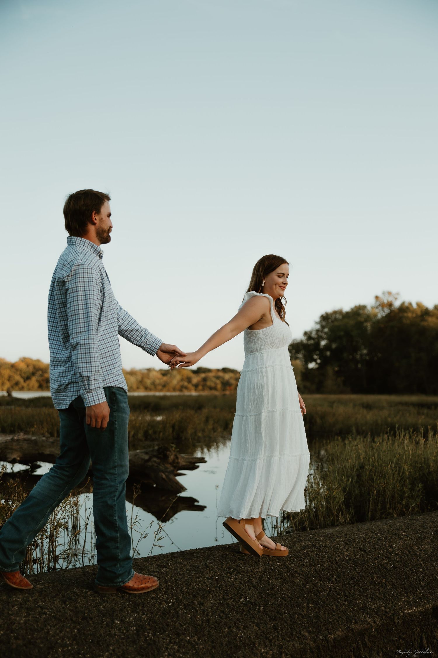A couple holds hands while walking along a riverbank at sunset, with her wearing a flowing white dress.