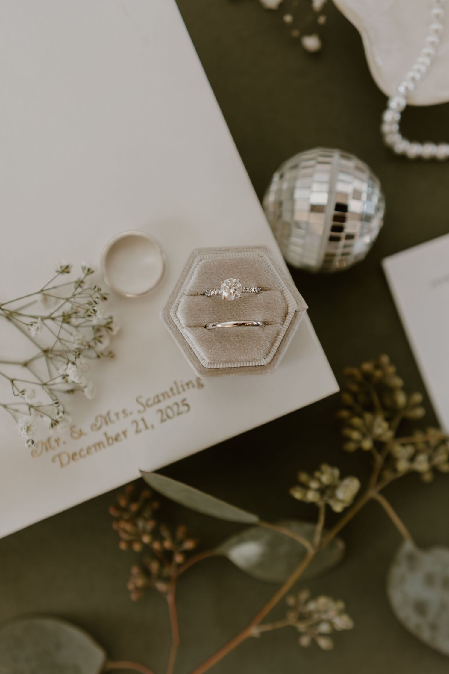 Wedding rings displayed in hexagonal box with dried flowers and disco ball decor on dark background.