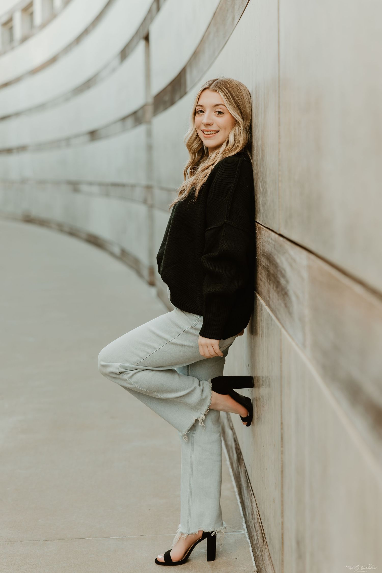 Woman in black sweater and light jeans poses against curved white wall.