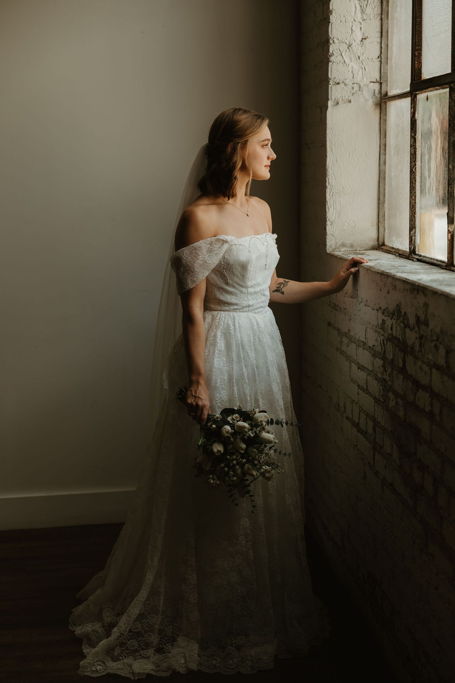 A bride in an off-shoulder lace wedding gown stands by a window holding a bouquet in moody natural lighting.