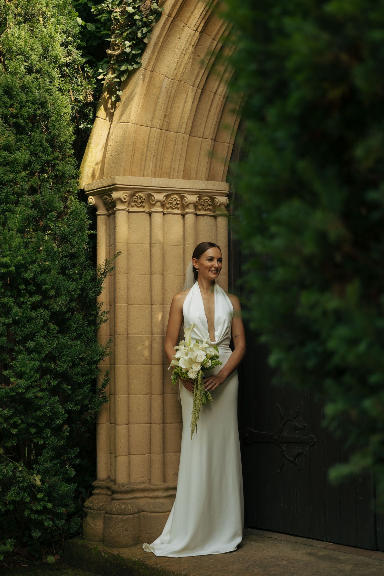 Bride in white halter gown holding bouquet stands under ornate stone arch surrounded by lush green foliage.