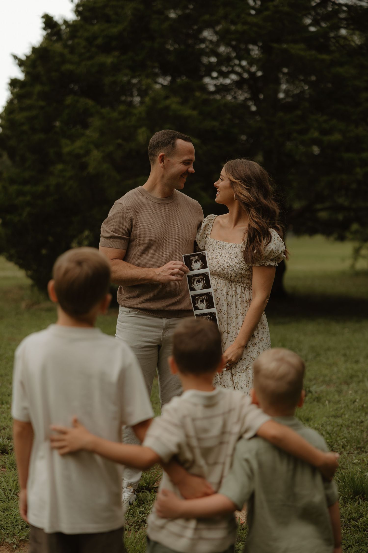 A couple smiles at each other holding an ultrasound photo outdoors while three young boys stand in the foreground.