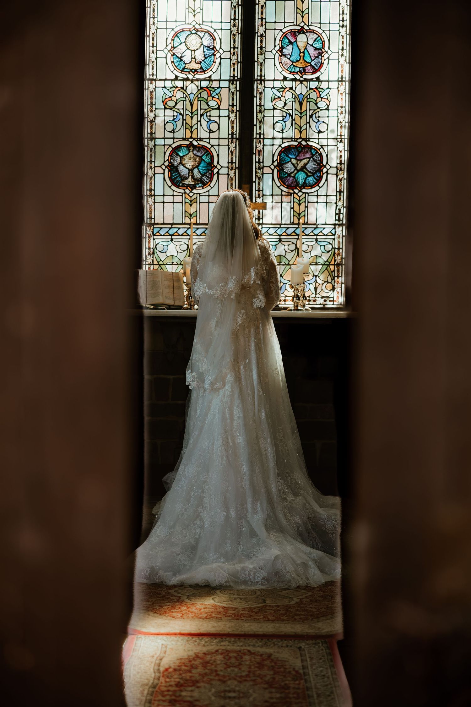 A white wedding dress silhouetted against ornate stained glass windows in a church, creating a dramatic backlit effect.