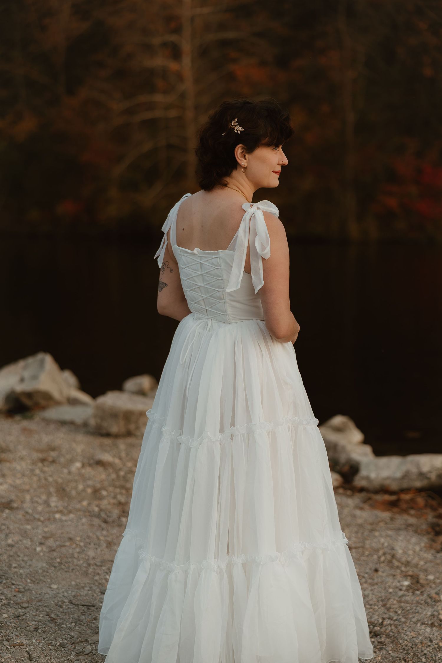 A child wearing a flowing white dress with ruffled layers stands on a gravel path during an autumn day.