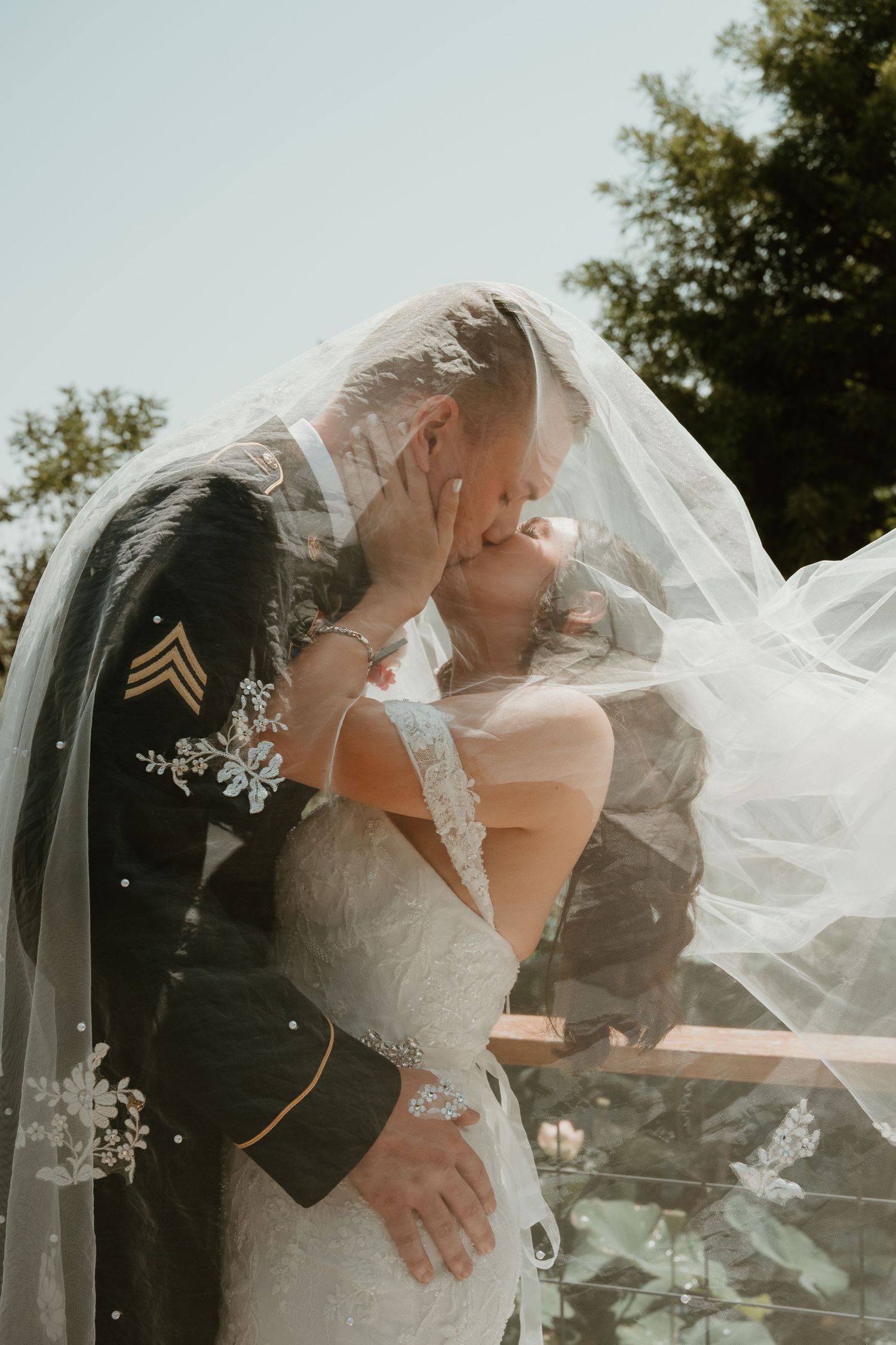 A romantic wedding couple embraces outdoors with sunlight filtering through a flowing white veil in the breeze.