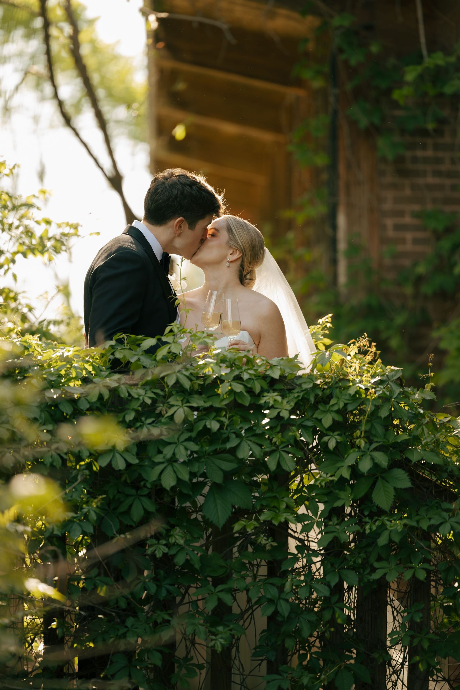 A bride and groom share a romantic kiss surrounded by lush green foliage outside a brick building on their wedding day.