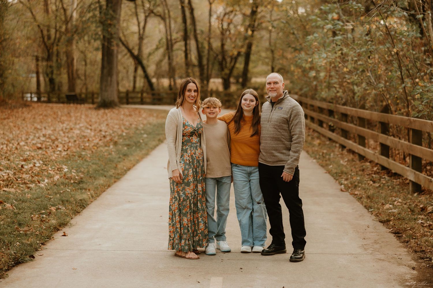 A family stands together on a paved path surrounded by autumn trees with fallen leaves on a cool fall day.
