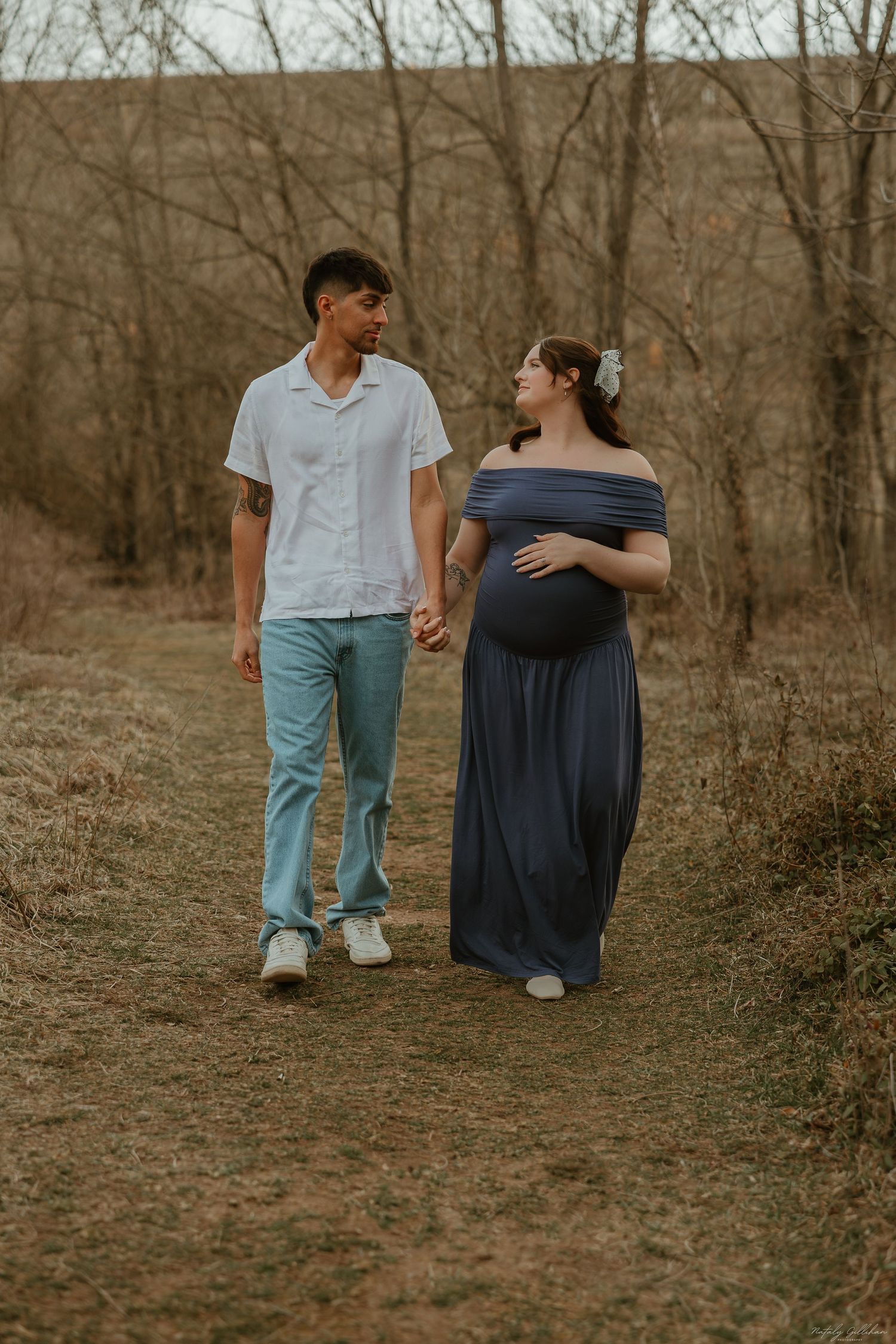 Expectant couple holds hands while walking on a dirt path through bare winter trees in warm earth tones.