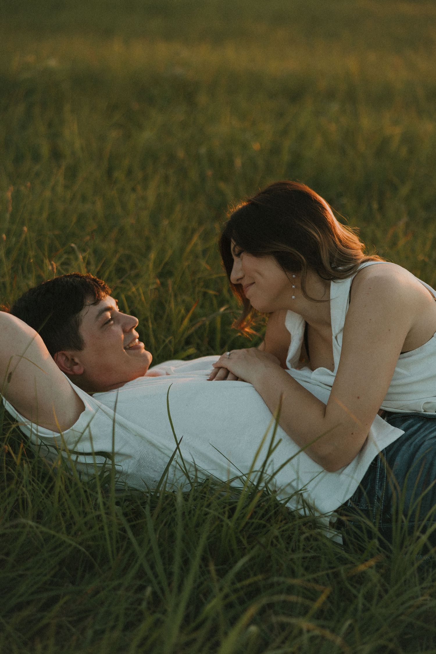Couple lying in green grass field, gazing at each other romantically during golden hour sunset.