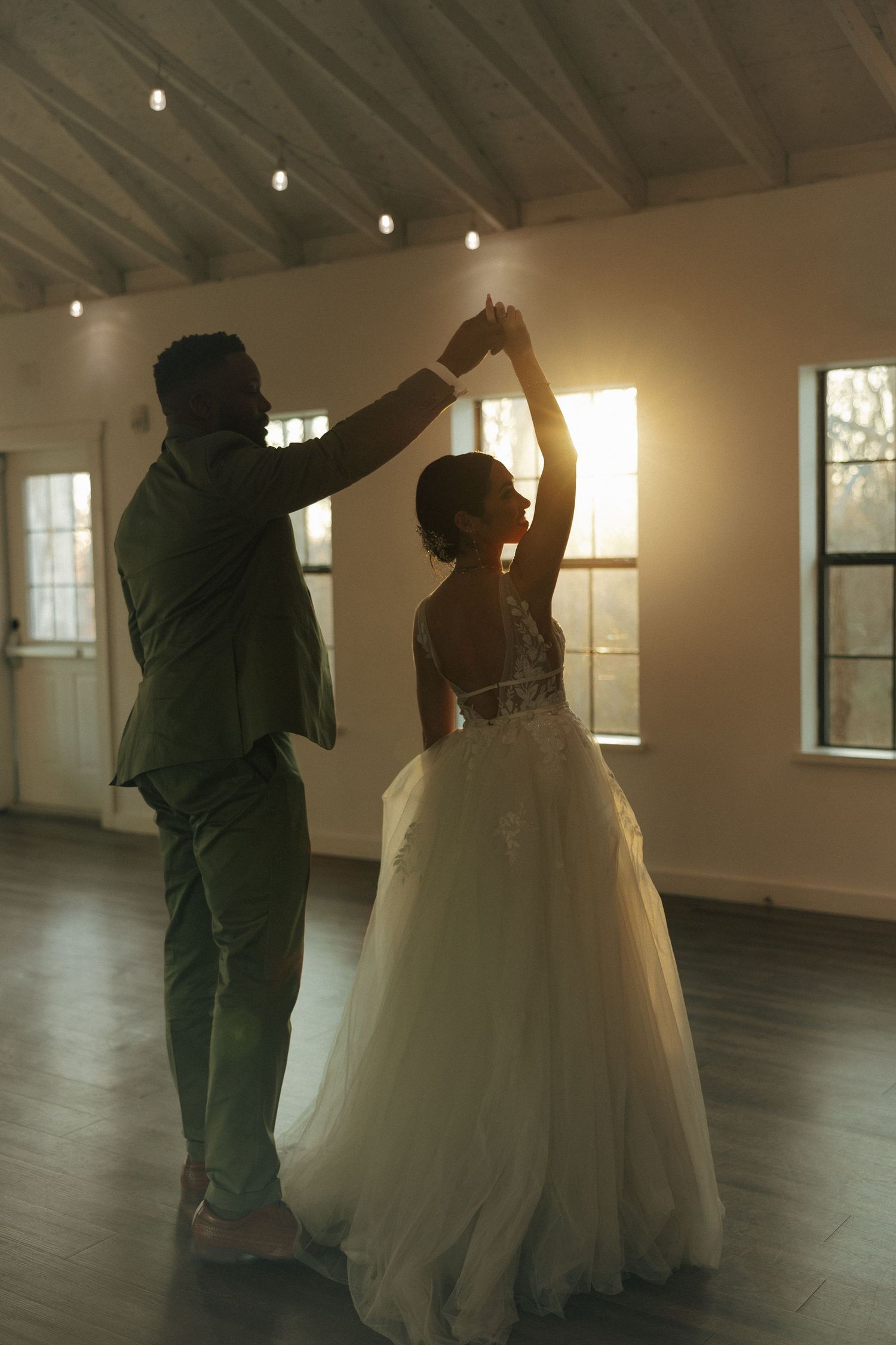 A couple shares an intimate dance together in a sunlit room with string lights overhead and a flowing white wedding gown.