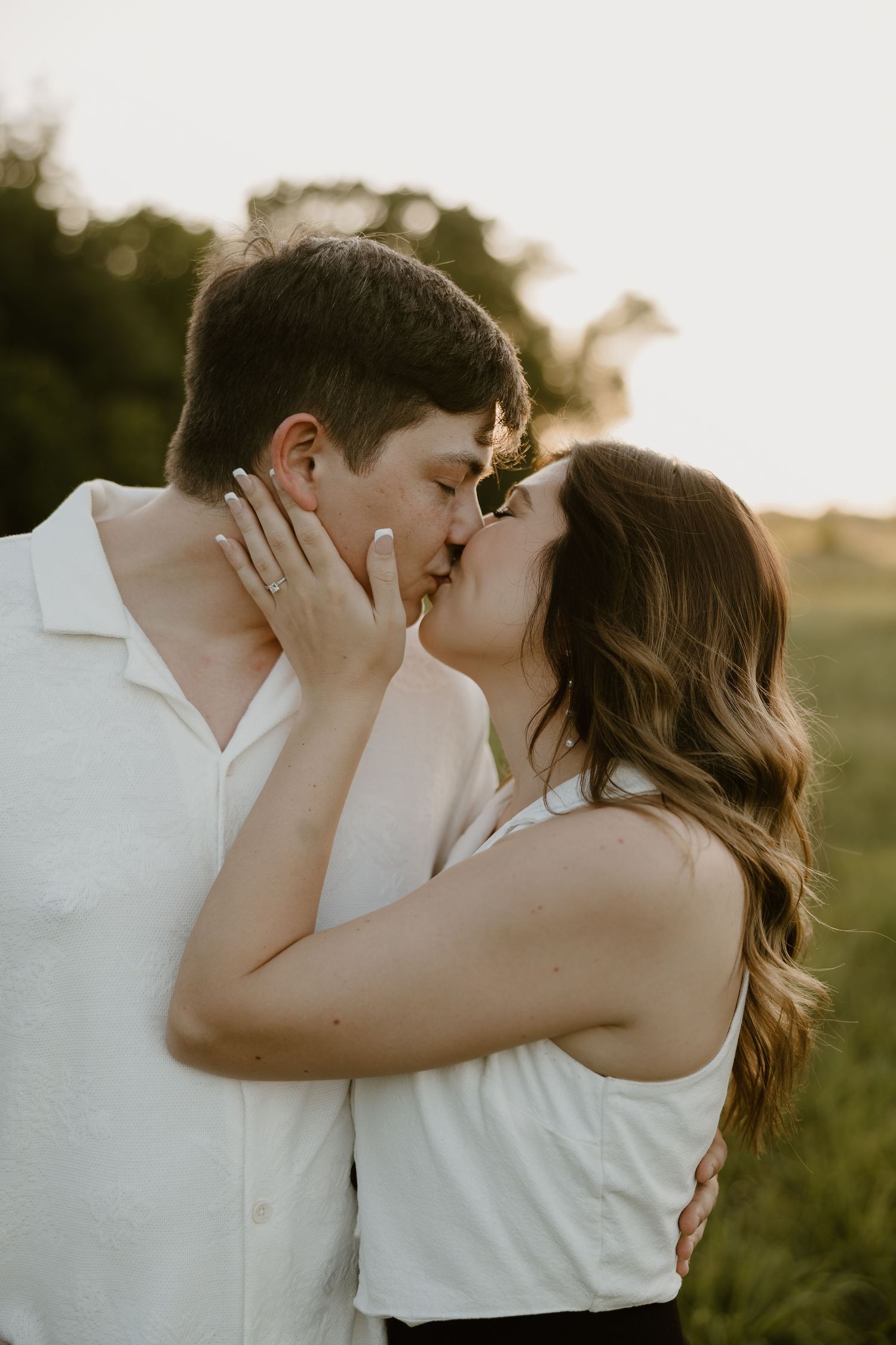A romantic couple shares a tender kiss in a sunlit field at golden hour, both wearing white tops.