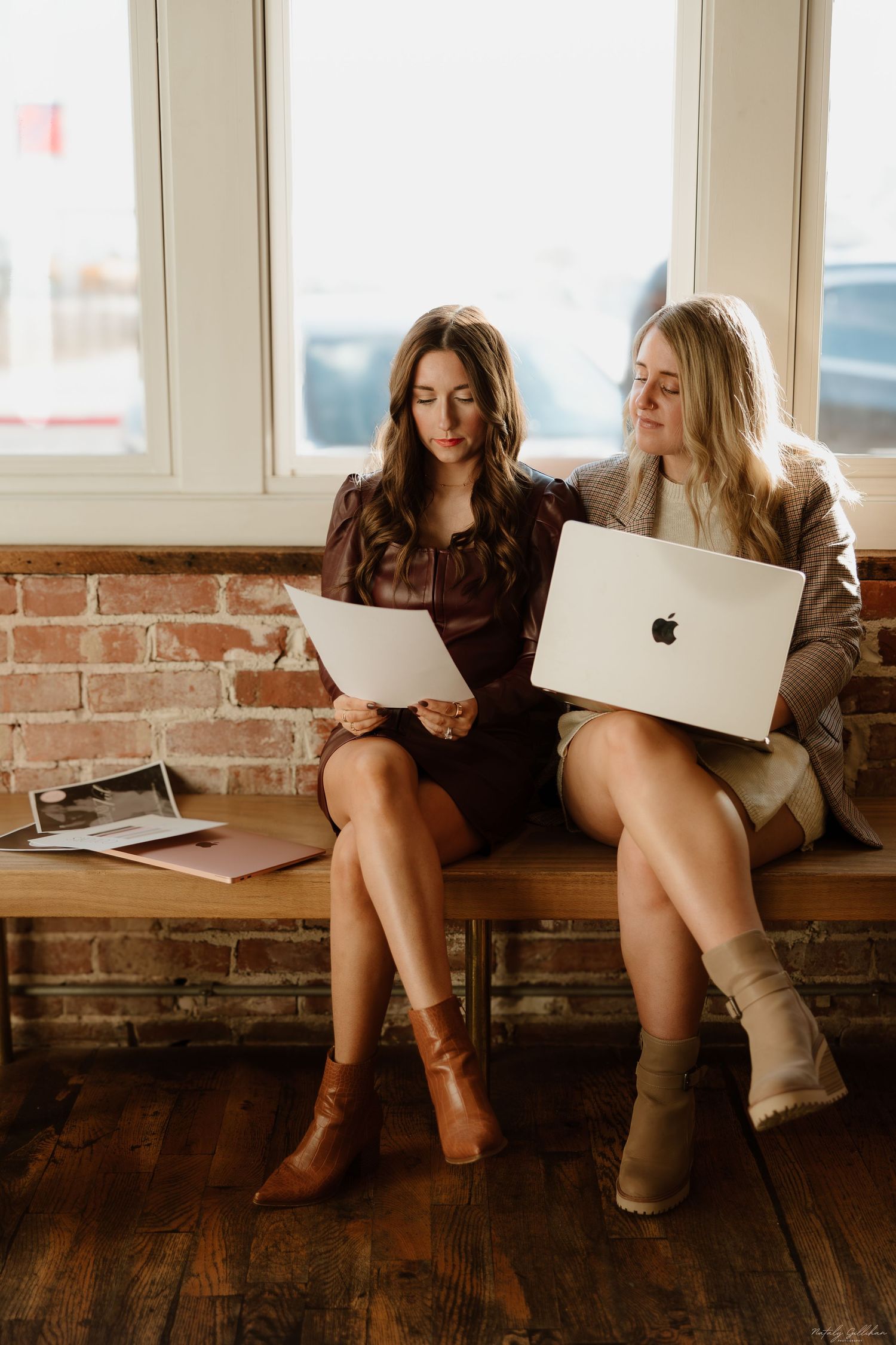 Two women in boots sit on a windowsill reviewing documents and working on a MacBook laptop in a rustic brick room.