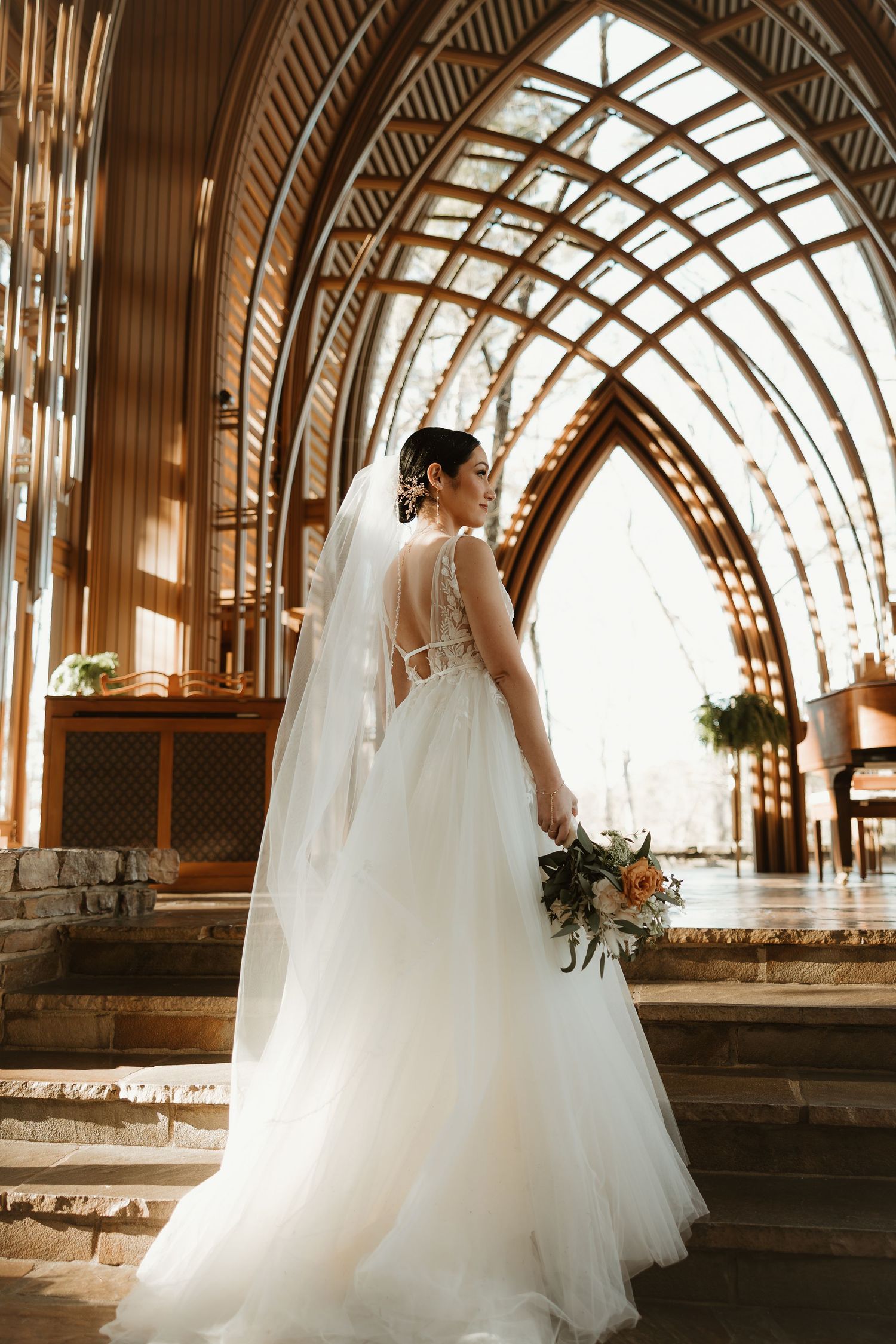 A bride in a flowing white tulle gown and veil stands in a chapel with dramatic arched windows and natural light.