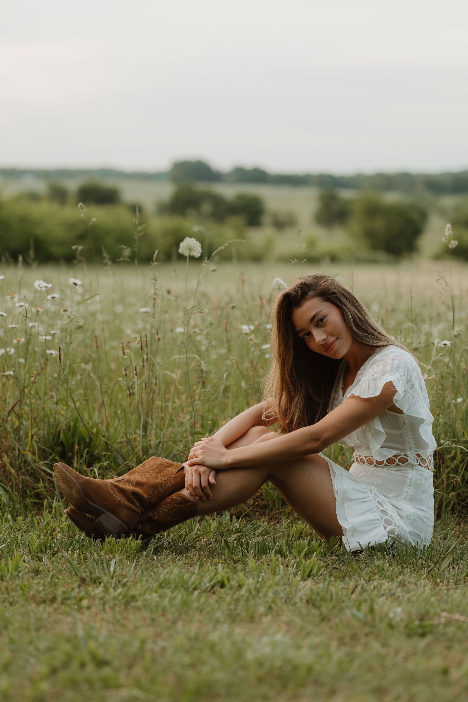 Young woman in white dress and brown cowboy boots sitting in a green meadow with wildflowers.