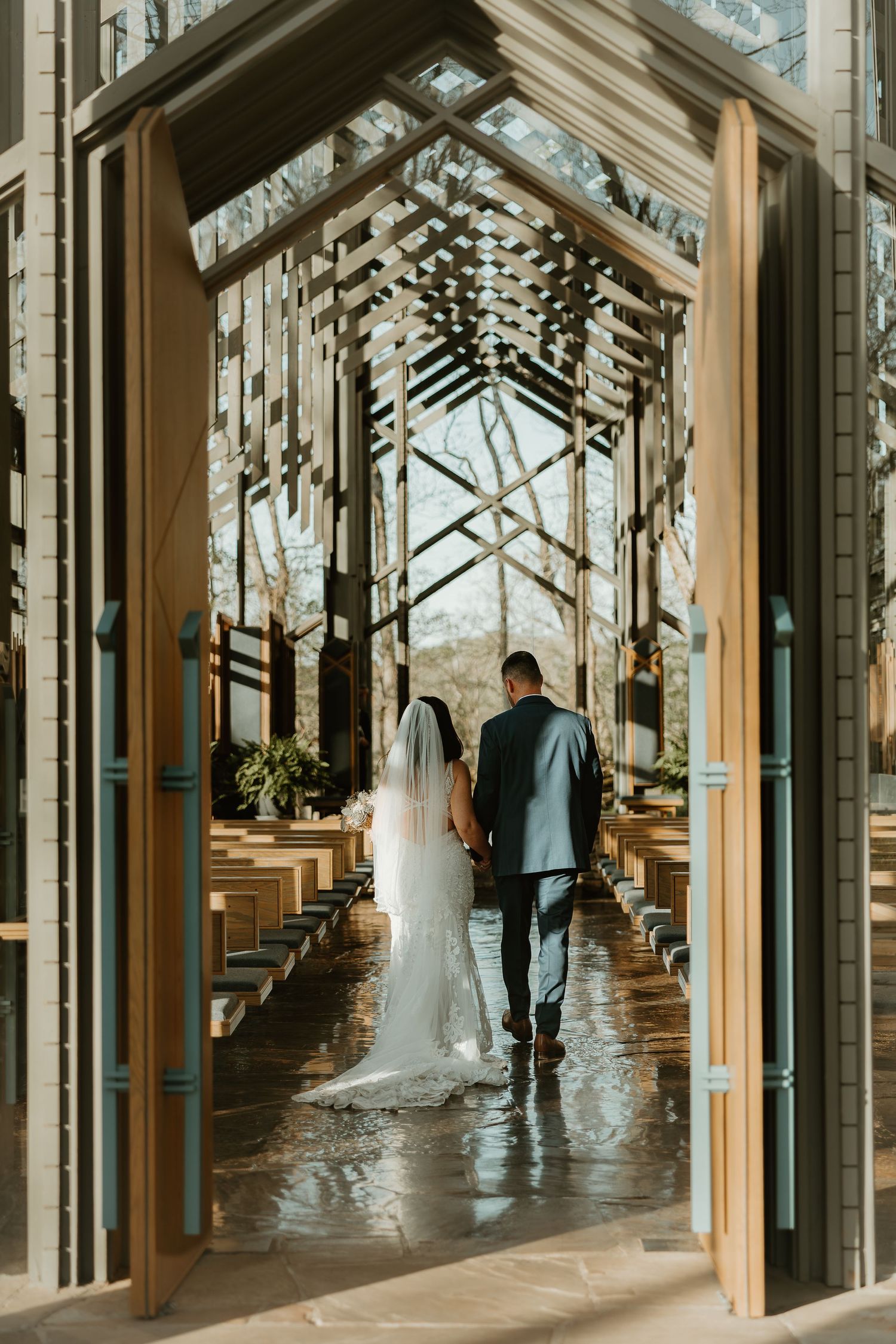 A couple walks through a glass chapel with wooden beams and sunlight streaming through the geometric architecture.