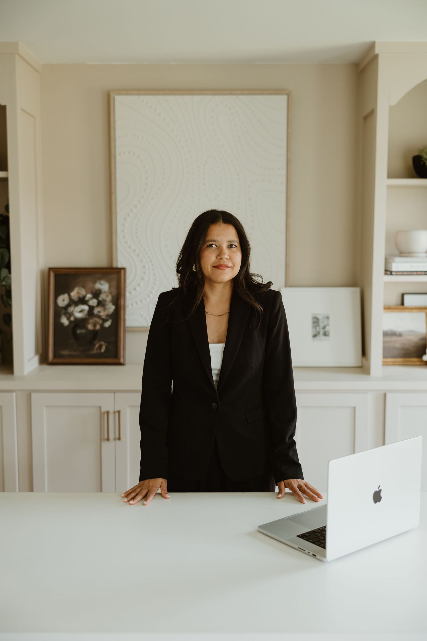 Professional standing at a white desk with laptop in modern home office with built-in shelving and artwork on display.