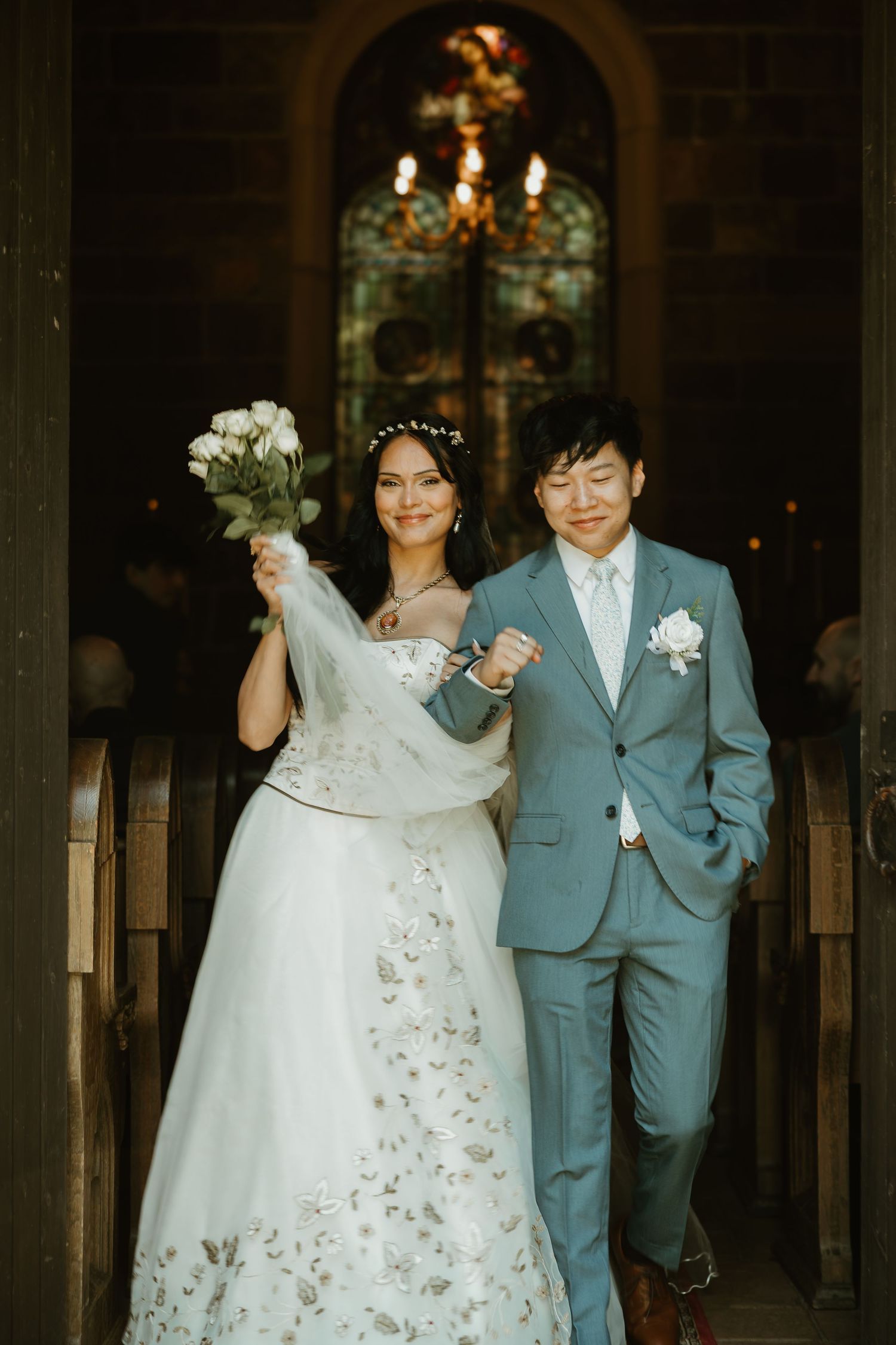 Newlywed couple walking down church aisle together, bride in white dress holding bouquet, groom in light blue suit.