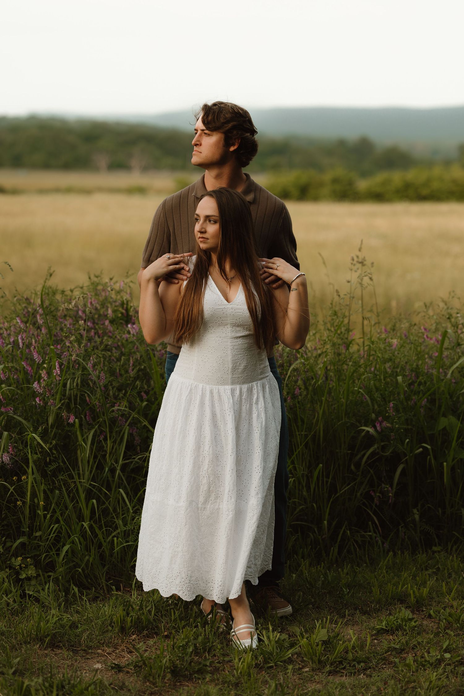 A couple stands in a lush green field, woman in white dress, man in brown shirt embracing her from behind.