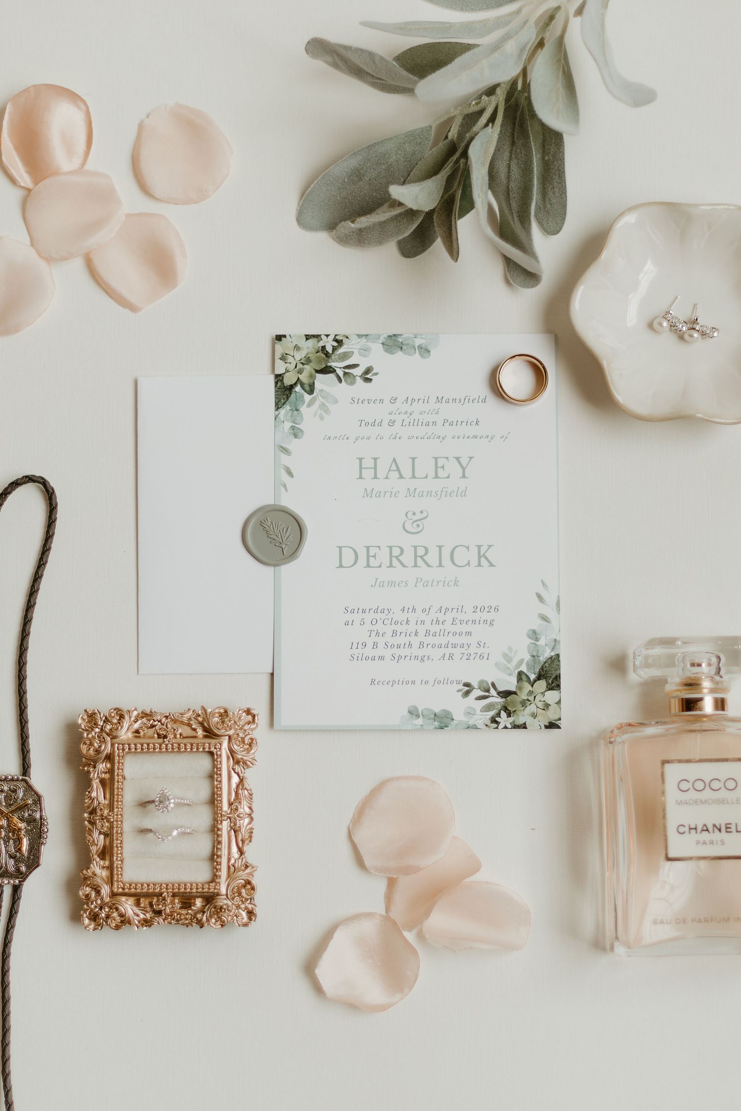 Elegant wedding flat lay featuring floral invitations for Haley and Derrick, gold rings, Chanel perfume, and rose petals.